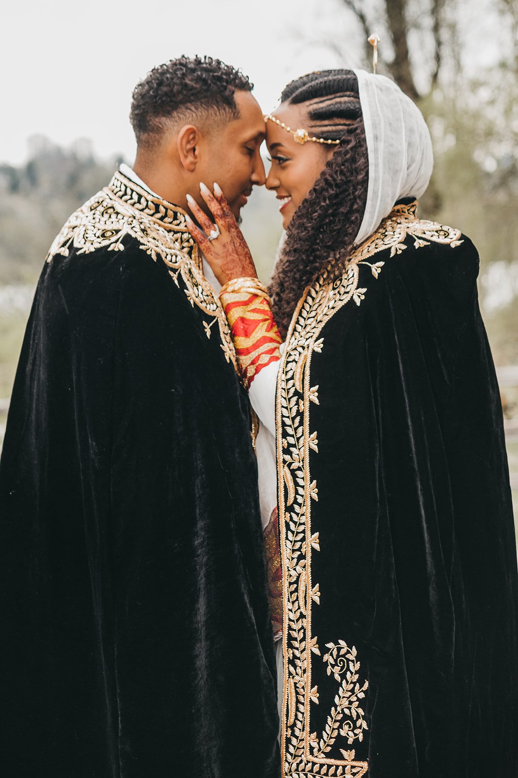 A couple stands close, dressed in matching ornate black capes with gold embroidery. The woman wears a headpiece and her hand, adorned with henna, rests on the man's shoulder. They appear happy and affectionate, with an outdoor setting in the backgrou