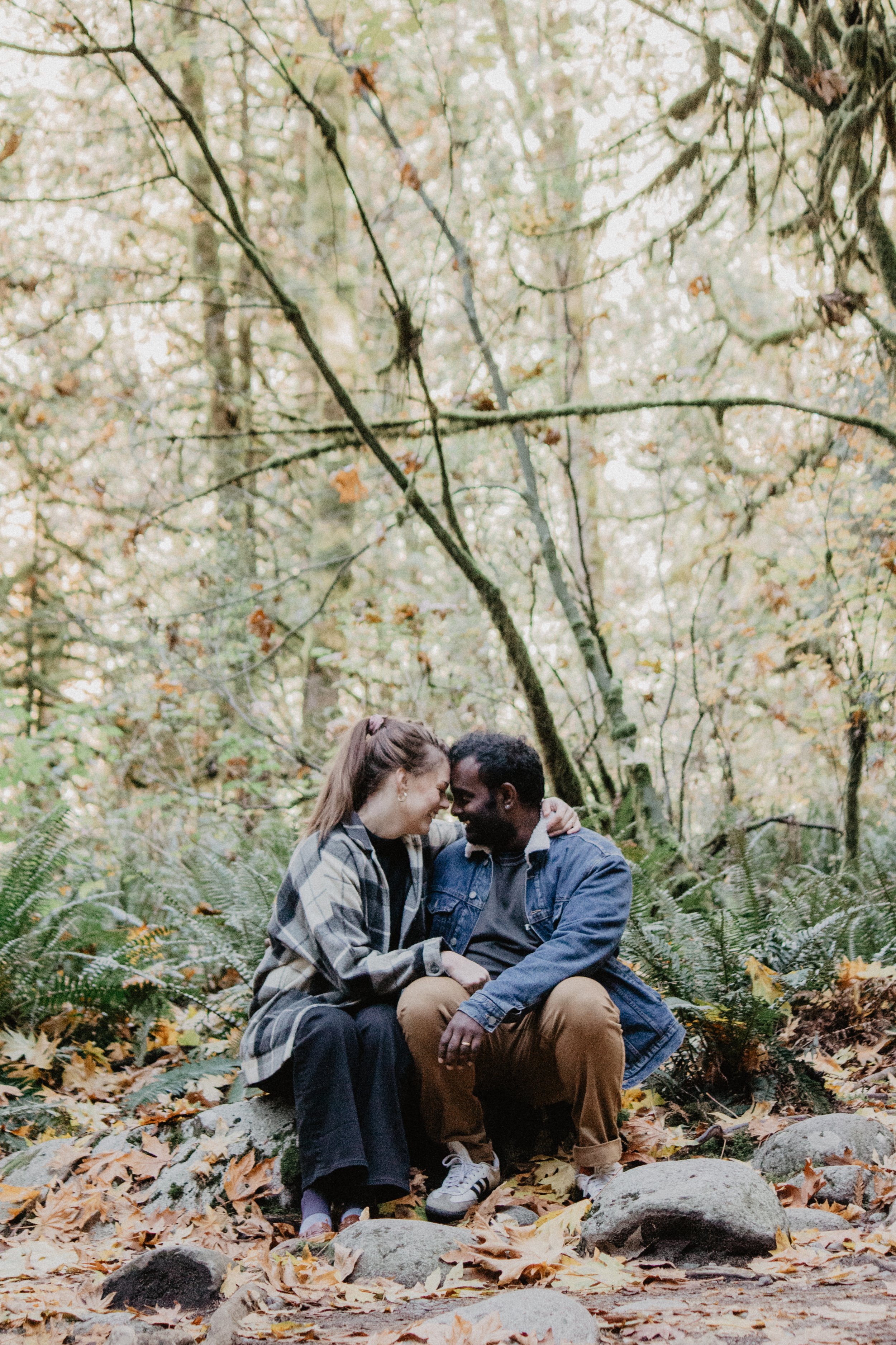 A couple sitting close together in a forest, surrounded by autumn leaves and trees.