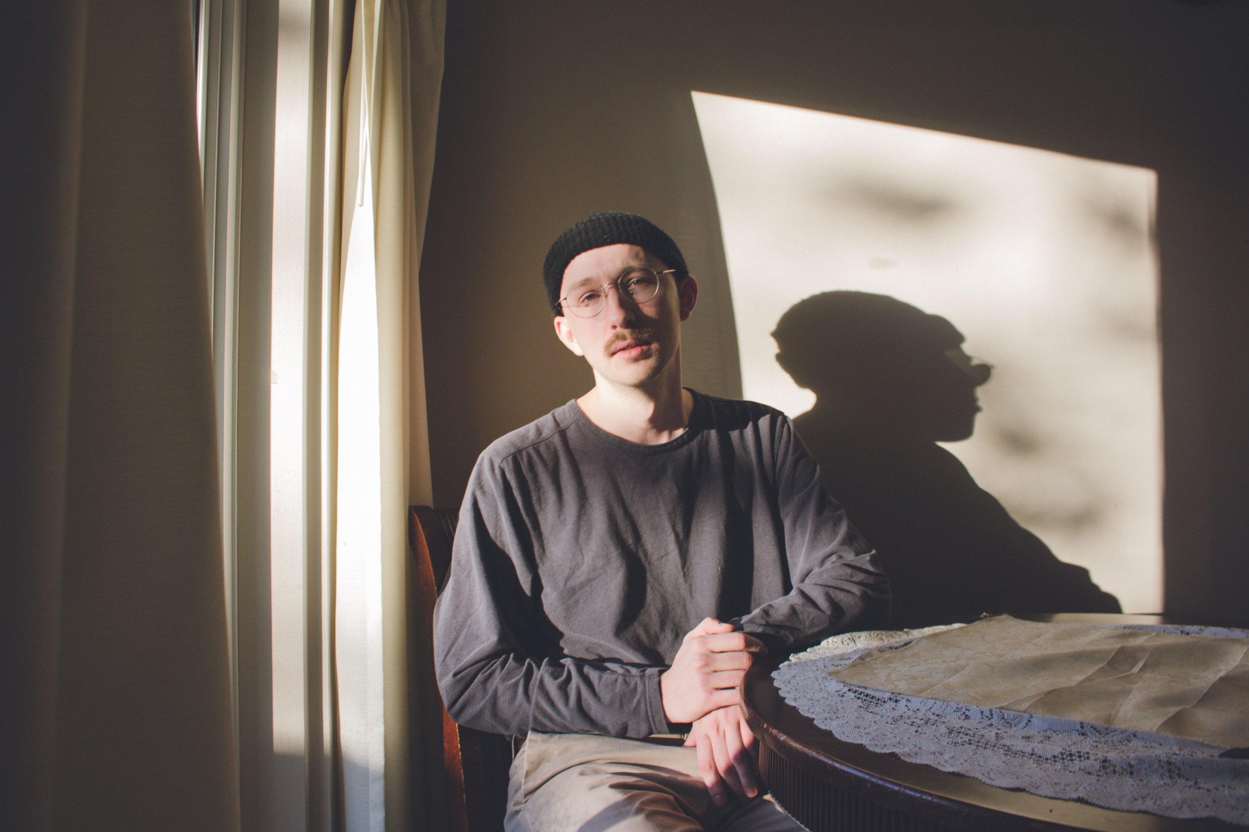 Person wearing glasses and a black beanie sitting by a table with sunlight casting a shadow on the wall.