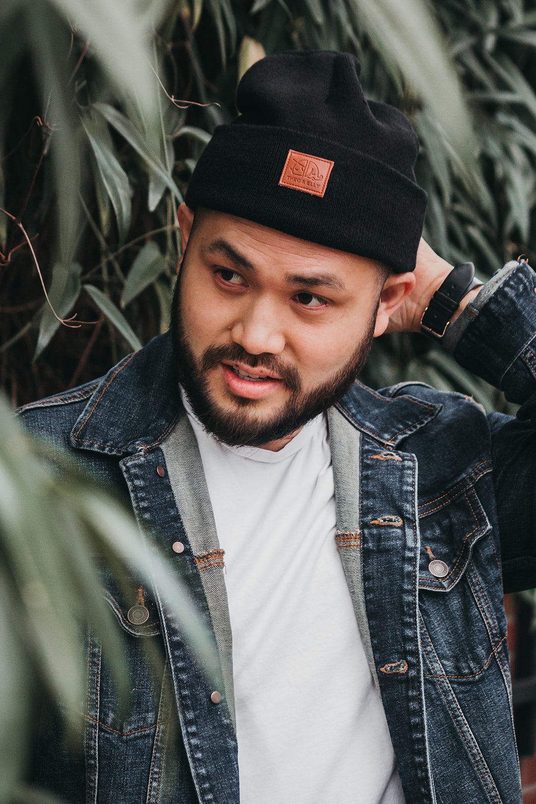 Man wearing a black beanie, denim jacket, and white shirt, with greenery in the background.