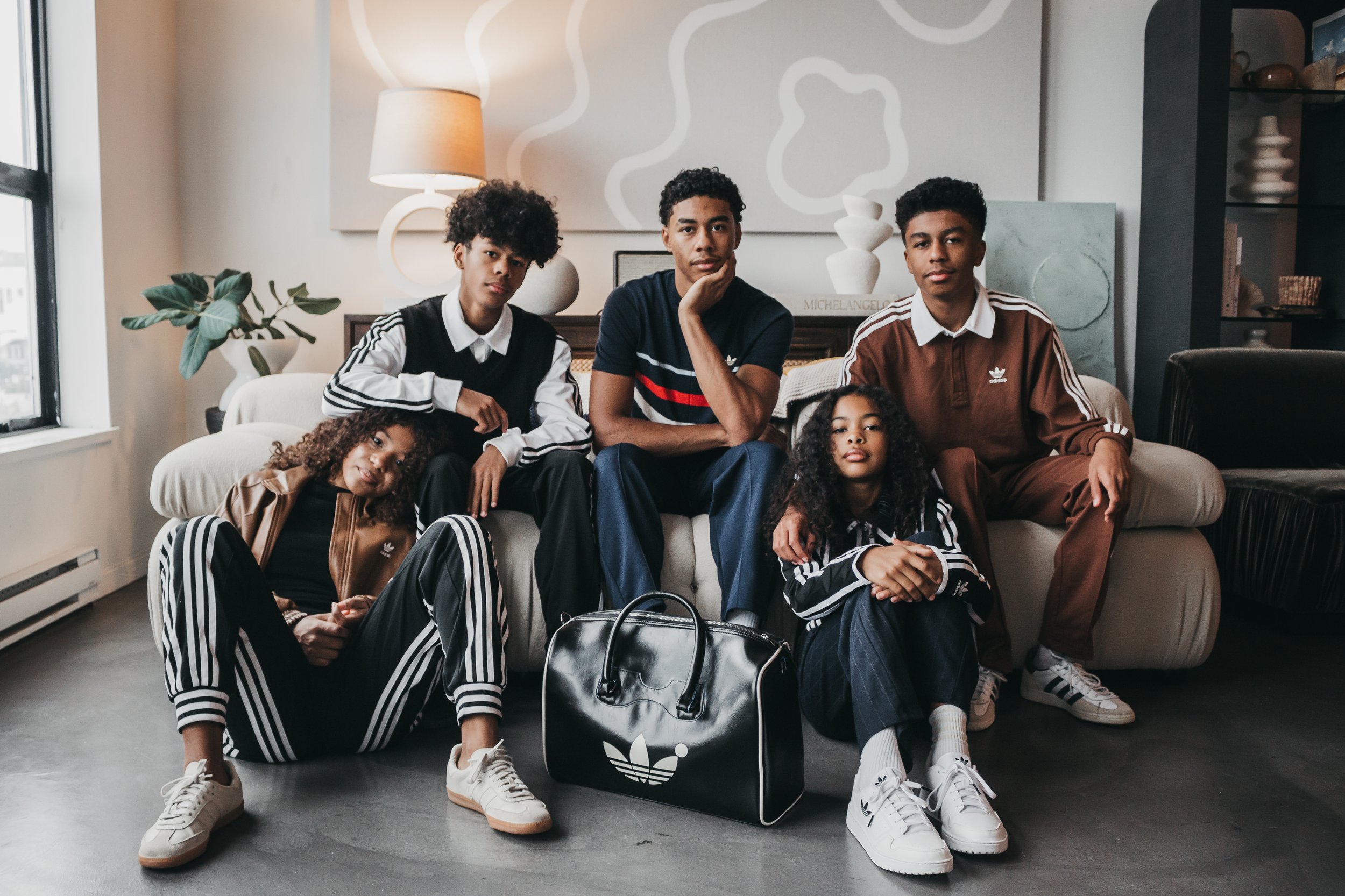 Group of five people with curly hair in casual Adidas outfits, sitting together in a cozy living room with modern decor, including a large lamp and abstract art on the wall.