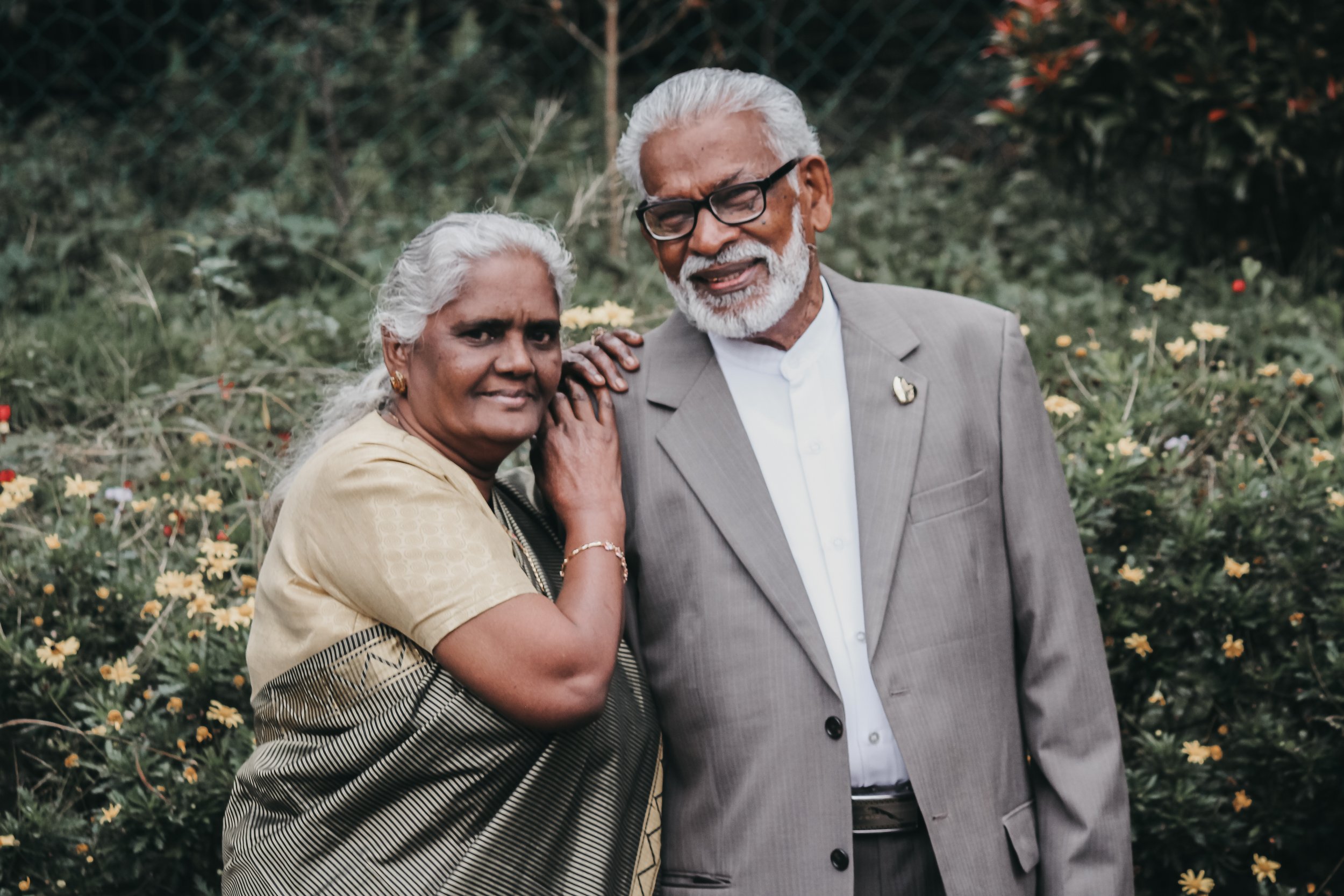 Elderly couple smiling outdoors, standing close with flowers and plants in the background.