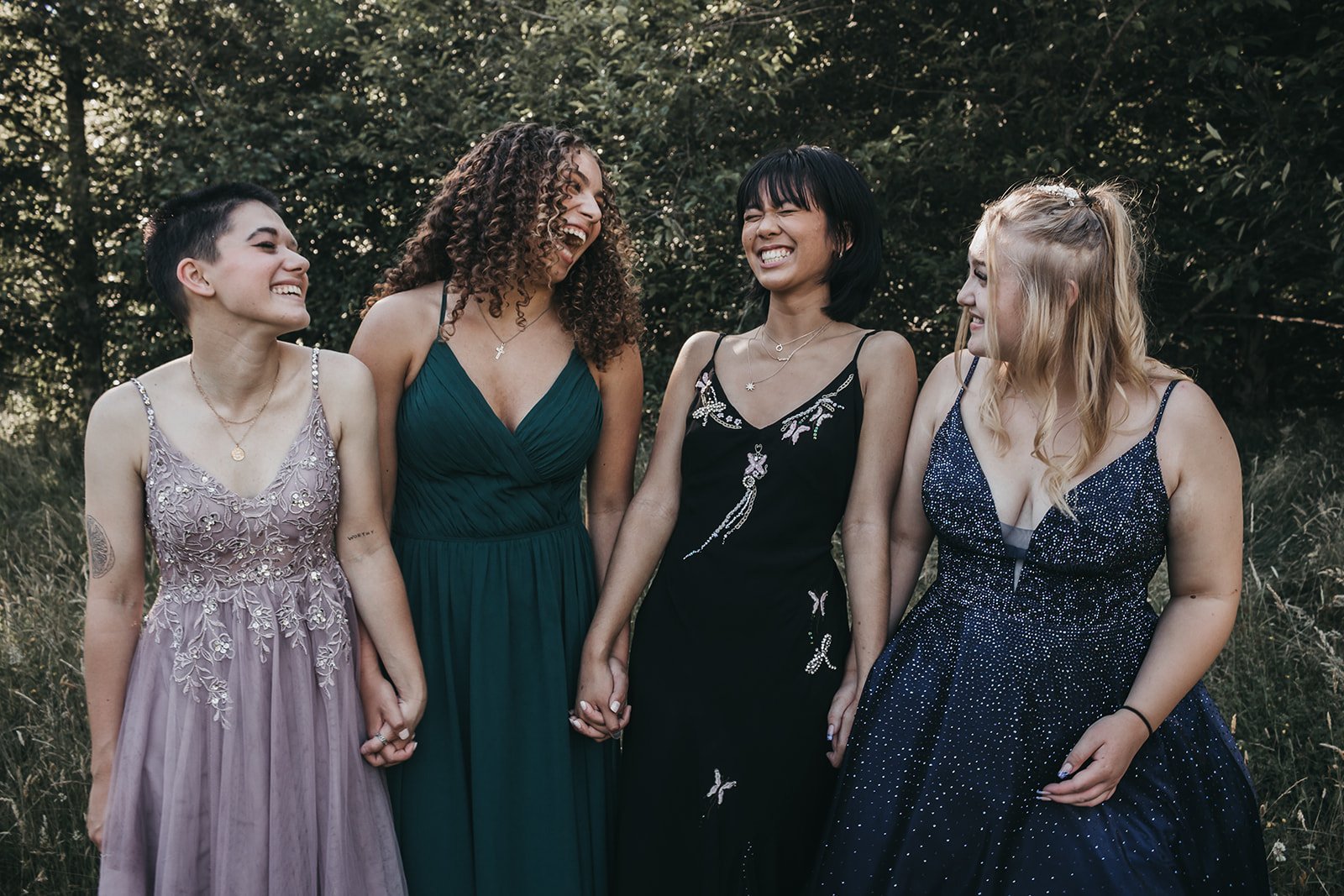 Four young women in formal dresses laughing and holding hands in a garden setting.