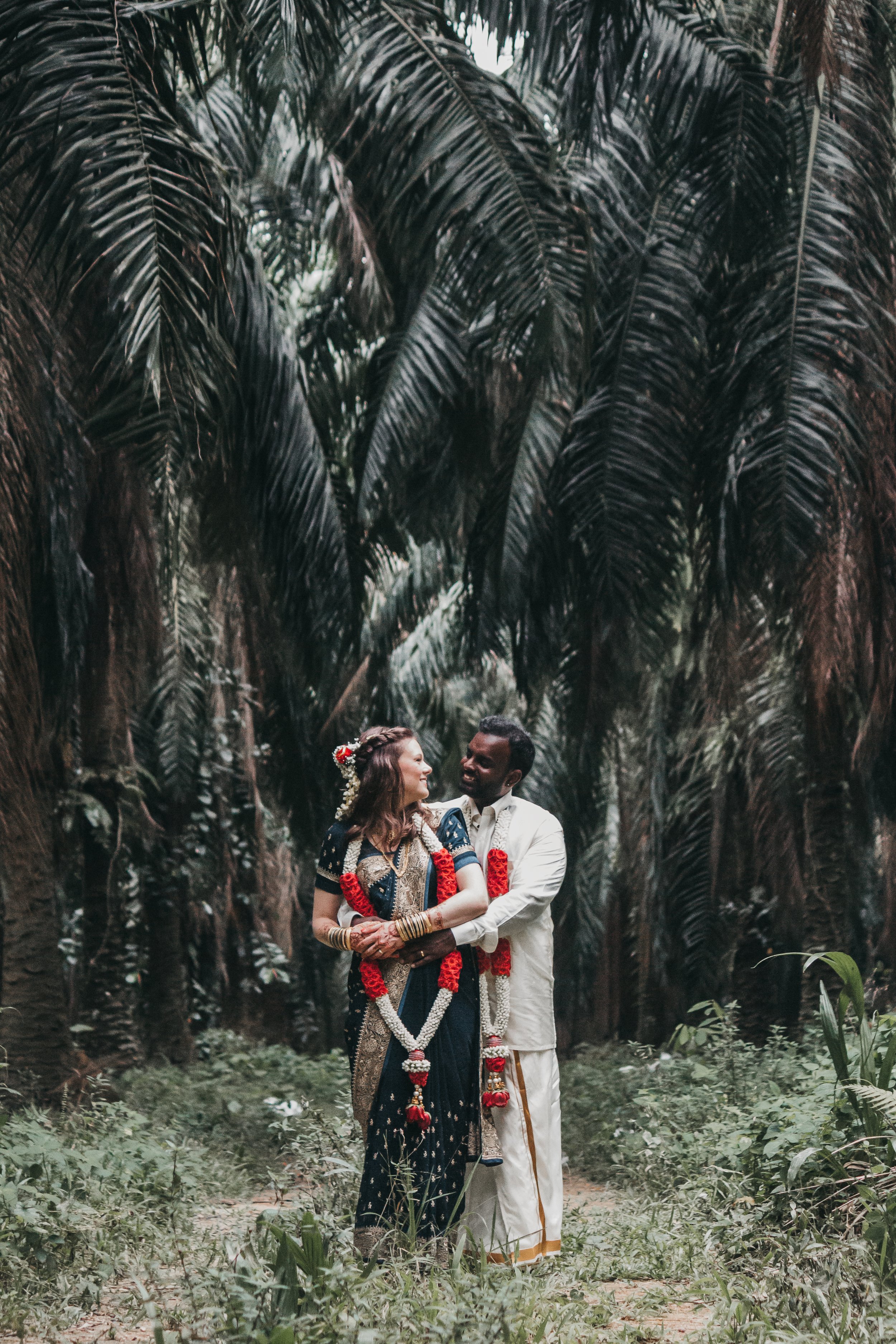 Couple in traditional attire standing in a lush forest, wearing floral garlands.