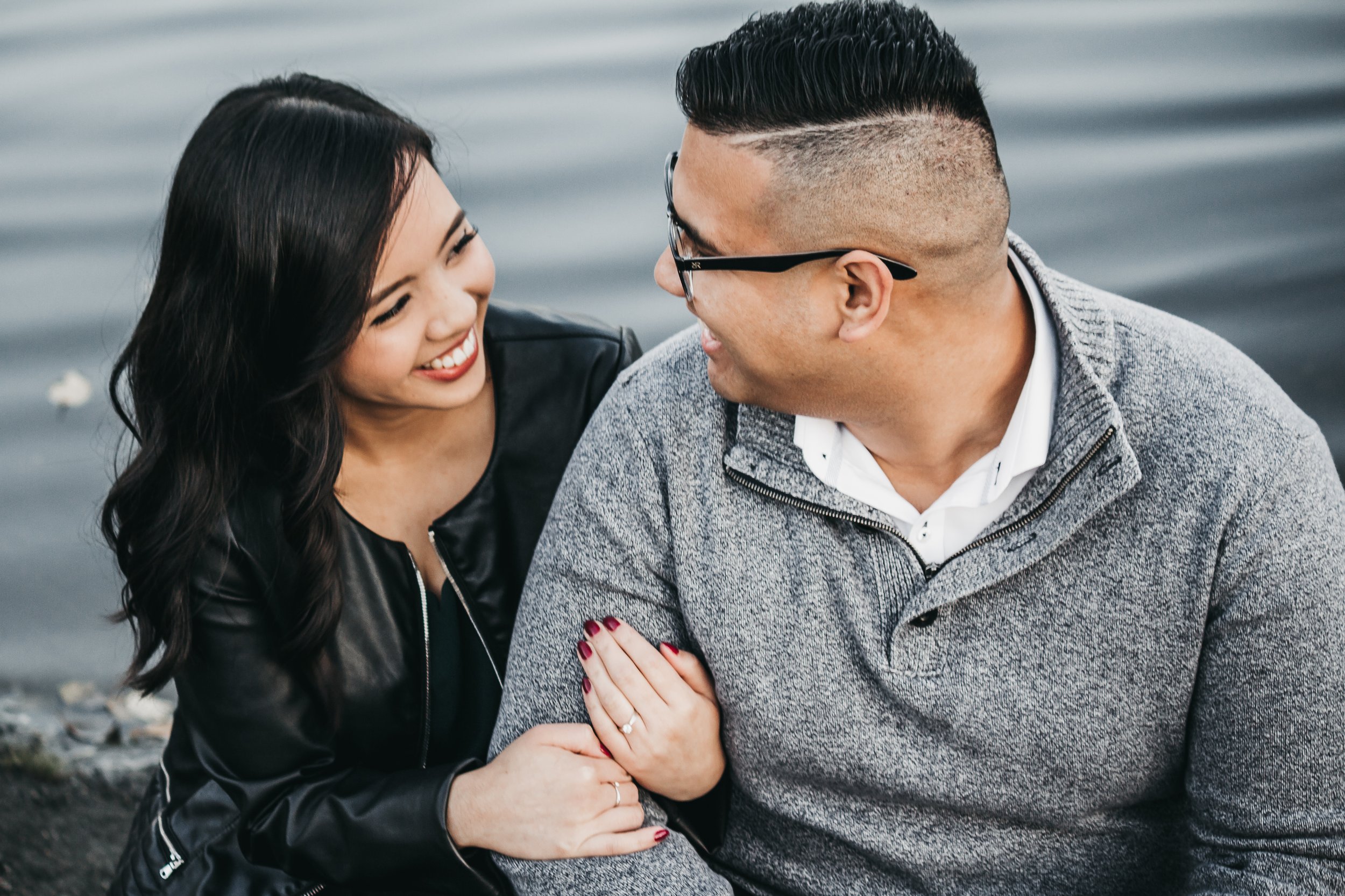Couple sitting by water, smiling at each other.