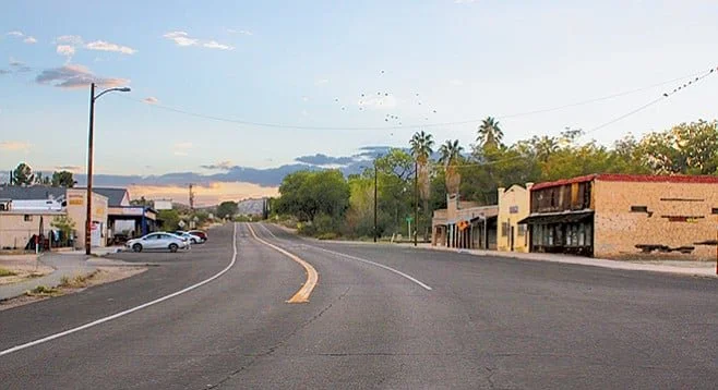 A small town street scene with a two-lane road, storefronts, parked cars, palm trees, and a sunset sky.