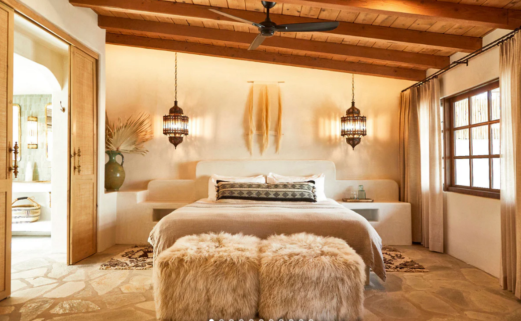 A cozy bedroom with a wooden ceiling, a large bed with white bedding and a black and white pillow, two hanging lantern-style lights, natural light coming through a large window with curtains, a fluffy ottoman at the foot of the bed, and a small nightstand with a glass of water.