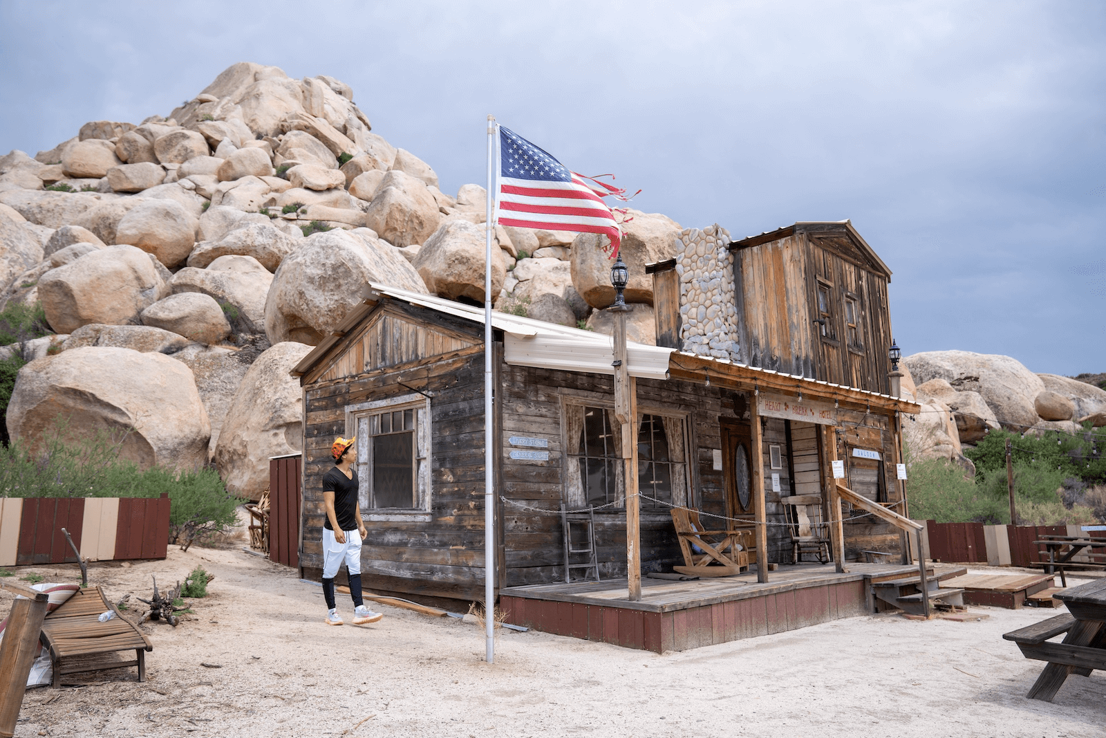 A woman in a black shirt and white pants standing outside a rustic wooden building with an American flag flying in front, set against a background of large rocks and cloudy sky.