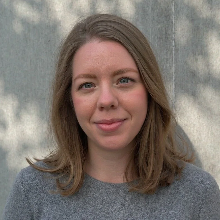 A woman with long brown hair wearing a gray sweater, standing against a textured wall with shadows.