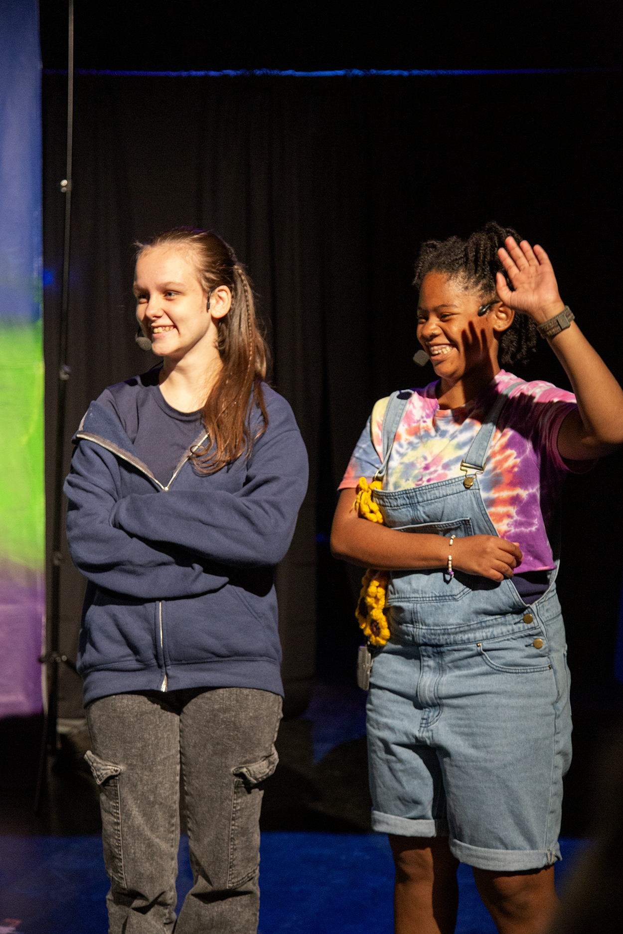 Two young women on stage, one with arms crossed in a blue hoodie and gray pants, the other with a smile, waving, in a tie-dye shirt and denim overalls, against a black curtain background.