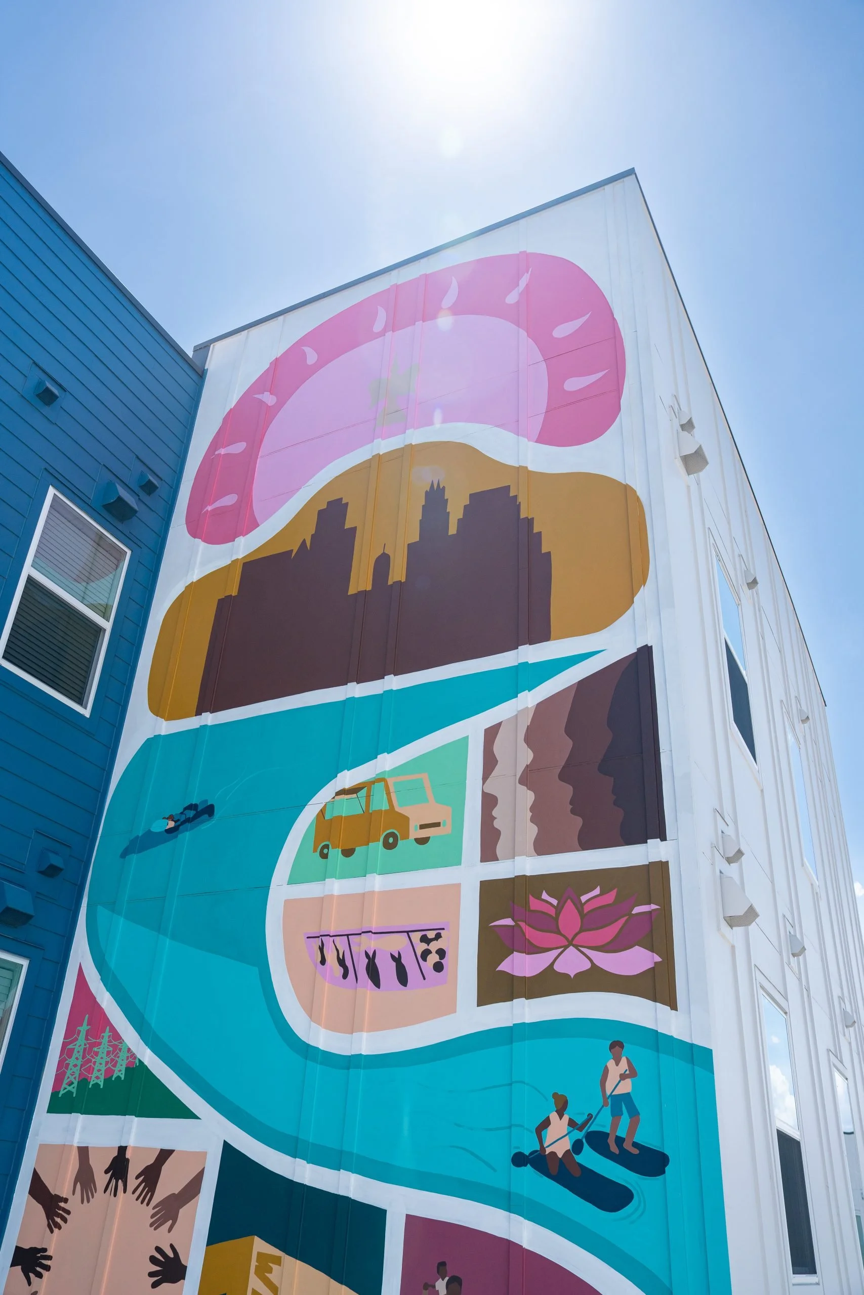 Colorful mural on the side of a building depicting various scenes including a pink donut, city skyline, a yellow car, gardening tools, a lotus flower, and people participating in outdoor activities against a clear blue sky.