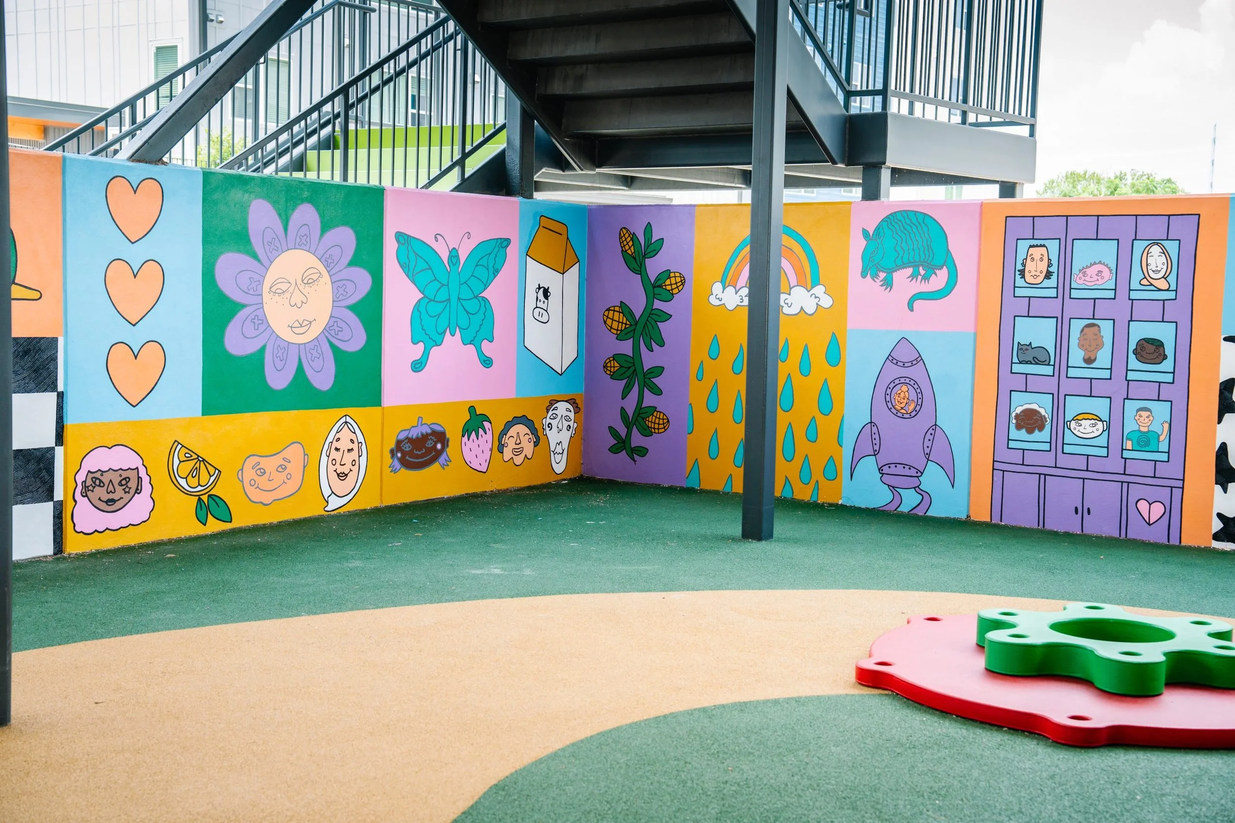Colorful playground wall featuring a mural with cartoon faces, animals, rainbows, and abstract designs. The ground is covered with multicolored rubber flooring. There are climbing structures and play equipment.