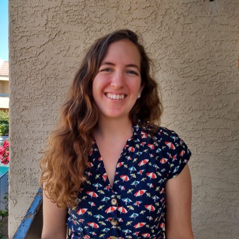 A young woman with long, wavy brown hair smiling in front of a beige textured wall, wearing a navy blue short-sleeved shirt with a colorful umbrella pattern.