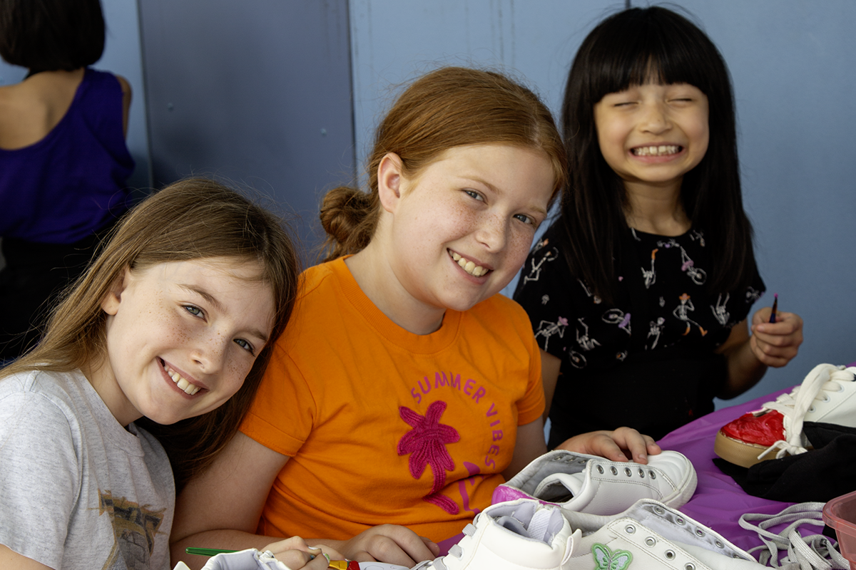 Three smiling young girls at a table with white sneakers on it, at an indoor event.