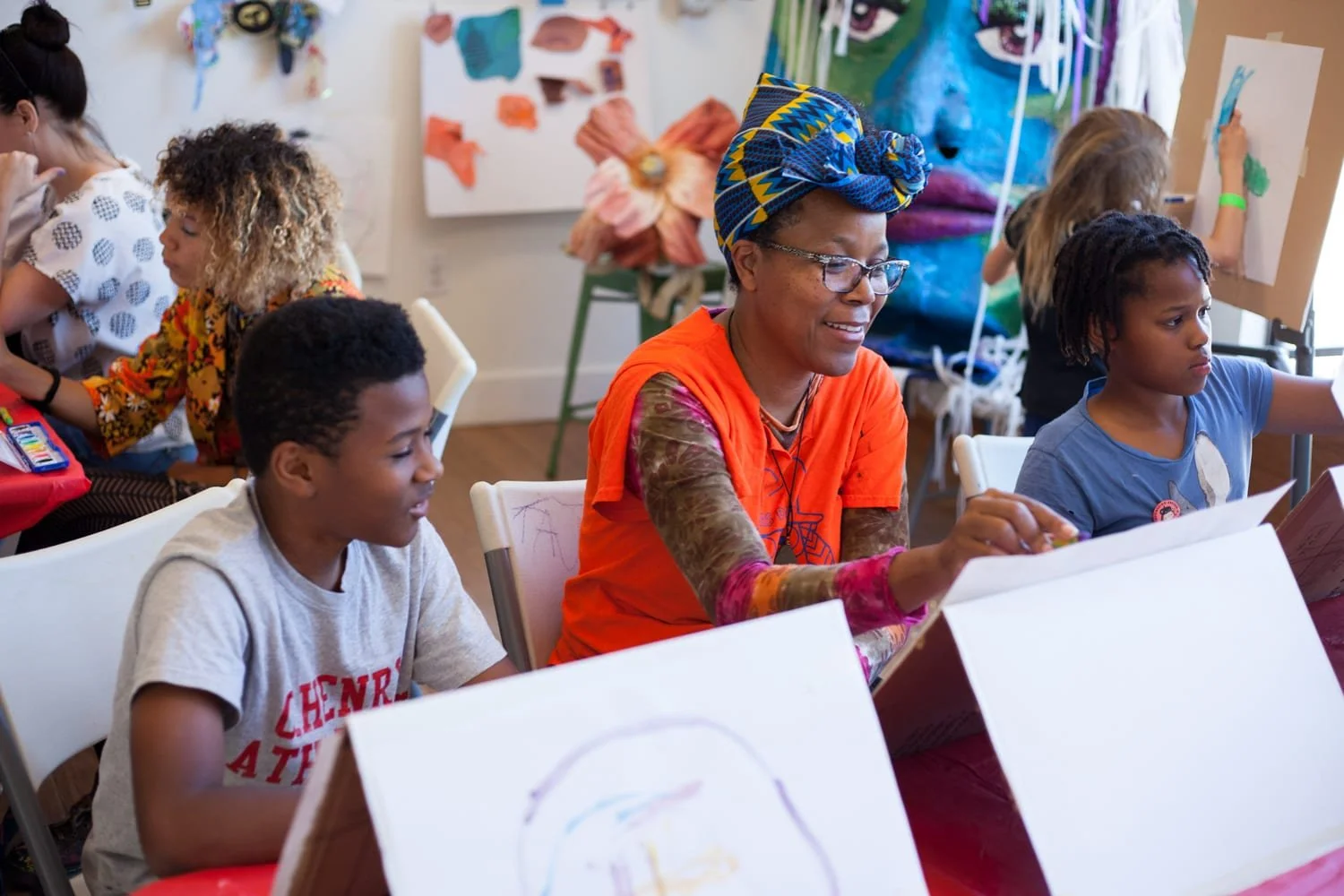 A diverse group of children and an adult woman sitting at tables, engaging in art activities such as drawing and painting, with colorful artwork and art supplies in the background.