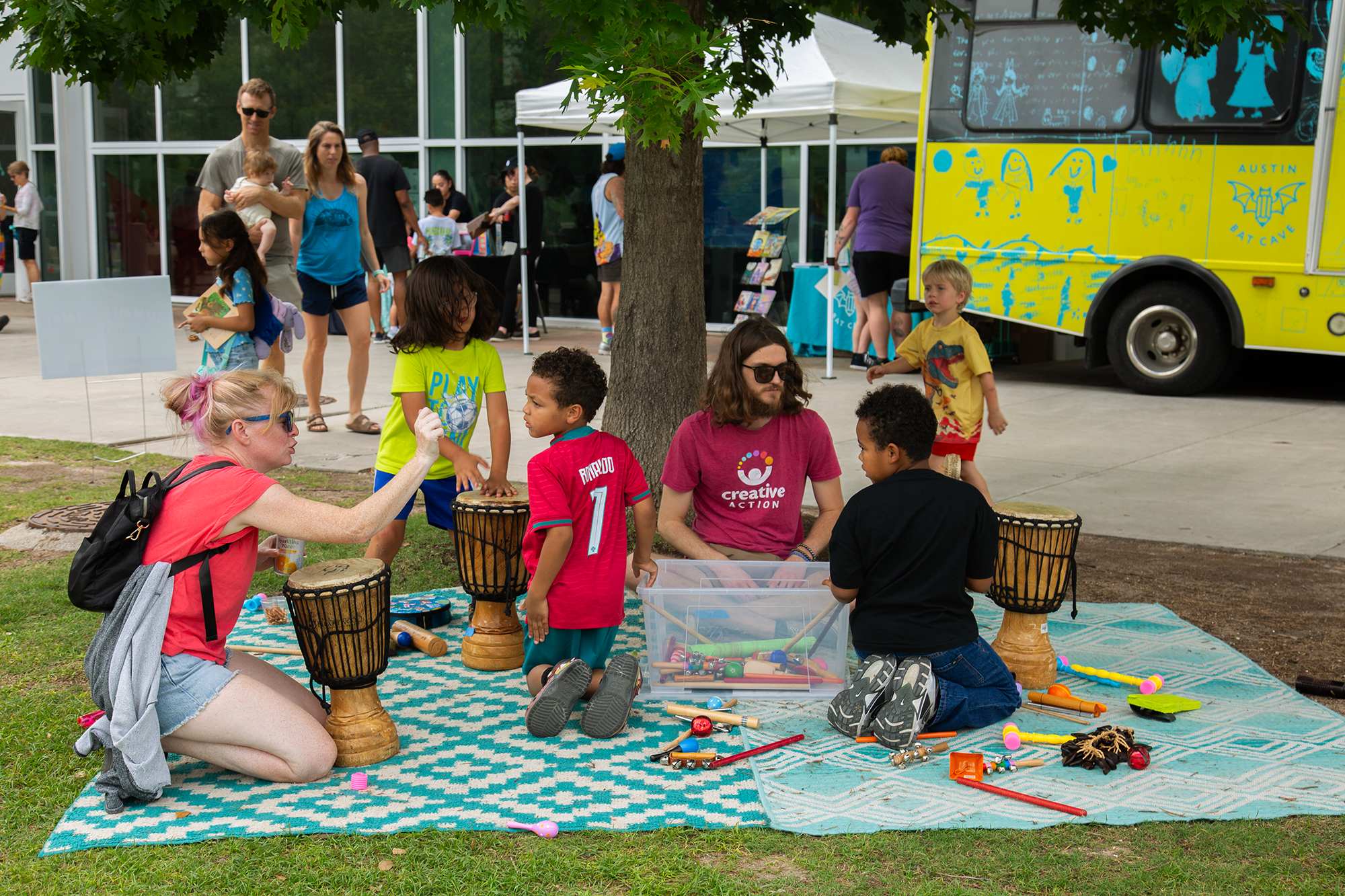 Children and adults participating in outdoor activities at a community event, with a blue and white striped rug, drums, and toys on the grass, and a yellow food truck labeled "Austin Bat Cave" nearby.
