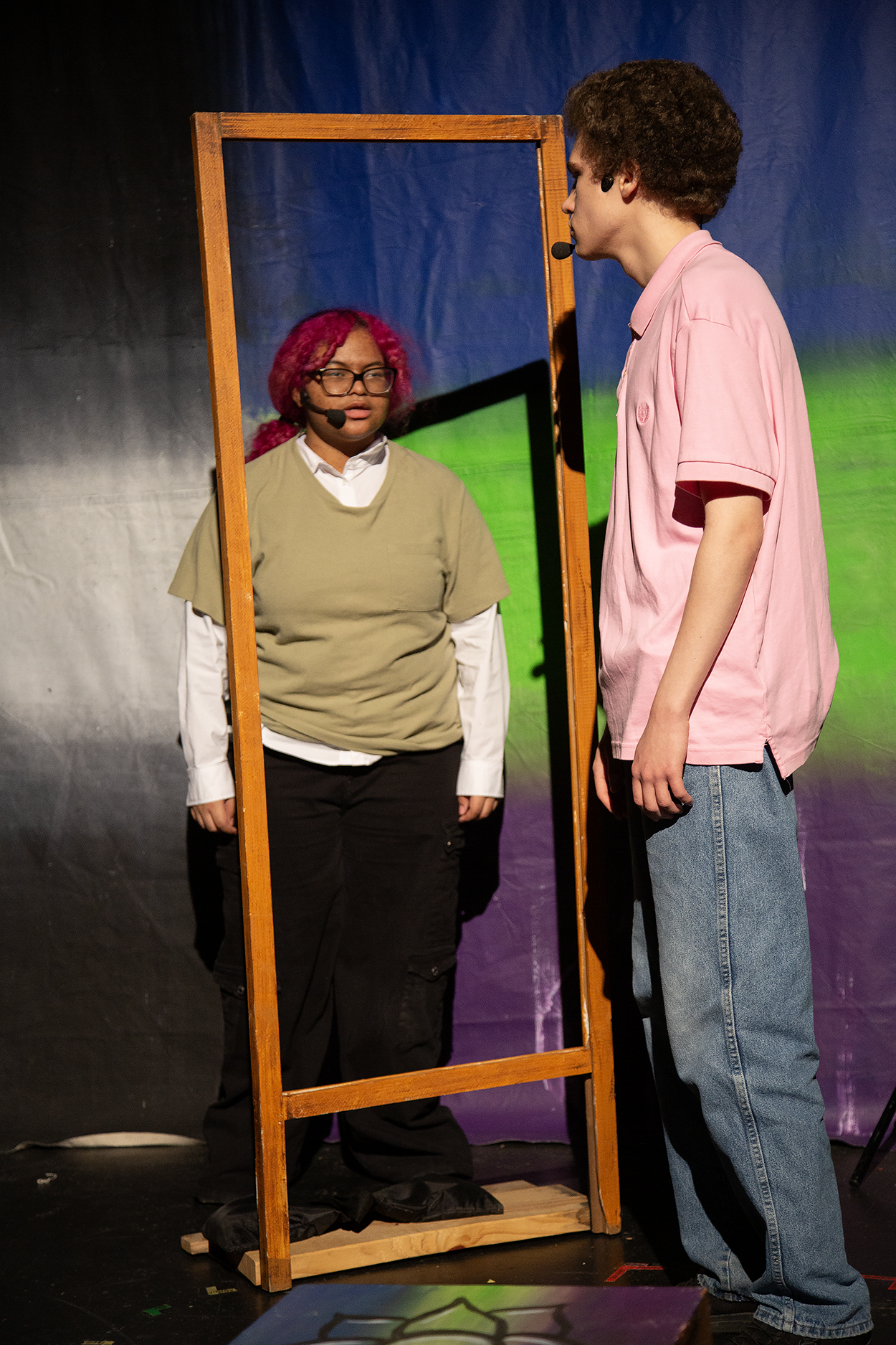 Two people on stage, one is looking at the other in a mirror. Both are wearing microphones. The person in the mirror has glasses, pink hair, a beige shirt, and black pants. The person outside the mirror has curly hair, a pink shirt, and blue jeans. The background is a colorful set with a blue and green backdrop.