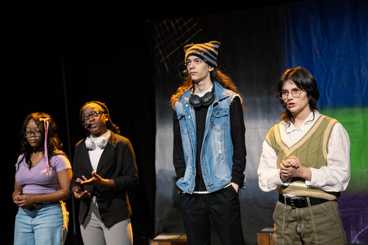 Four young people stand on stage with a black background, wearing casual clothes and headsets, possibly rehearsing for a performance.