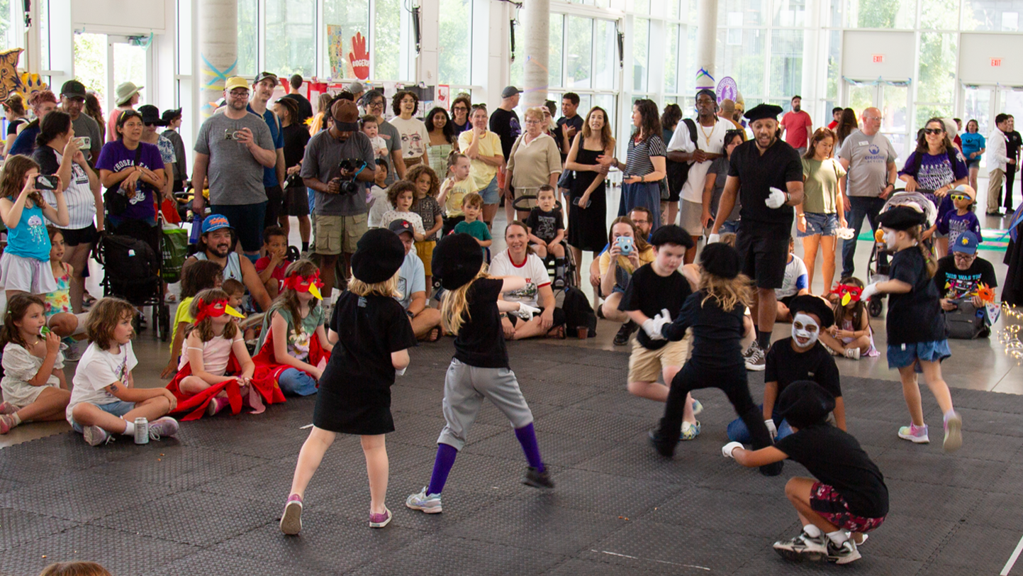 Children dressed as puppets perform a puppet show while an audience of children and adults watches inside a bright, spacious room with large windows.