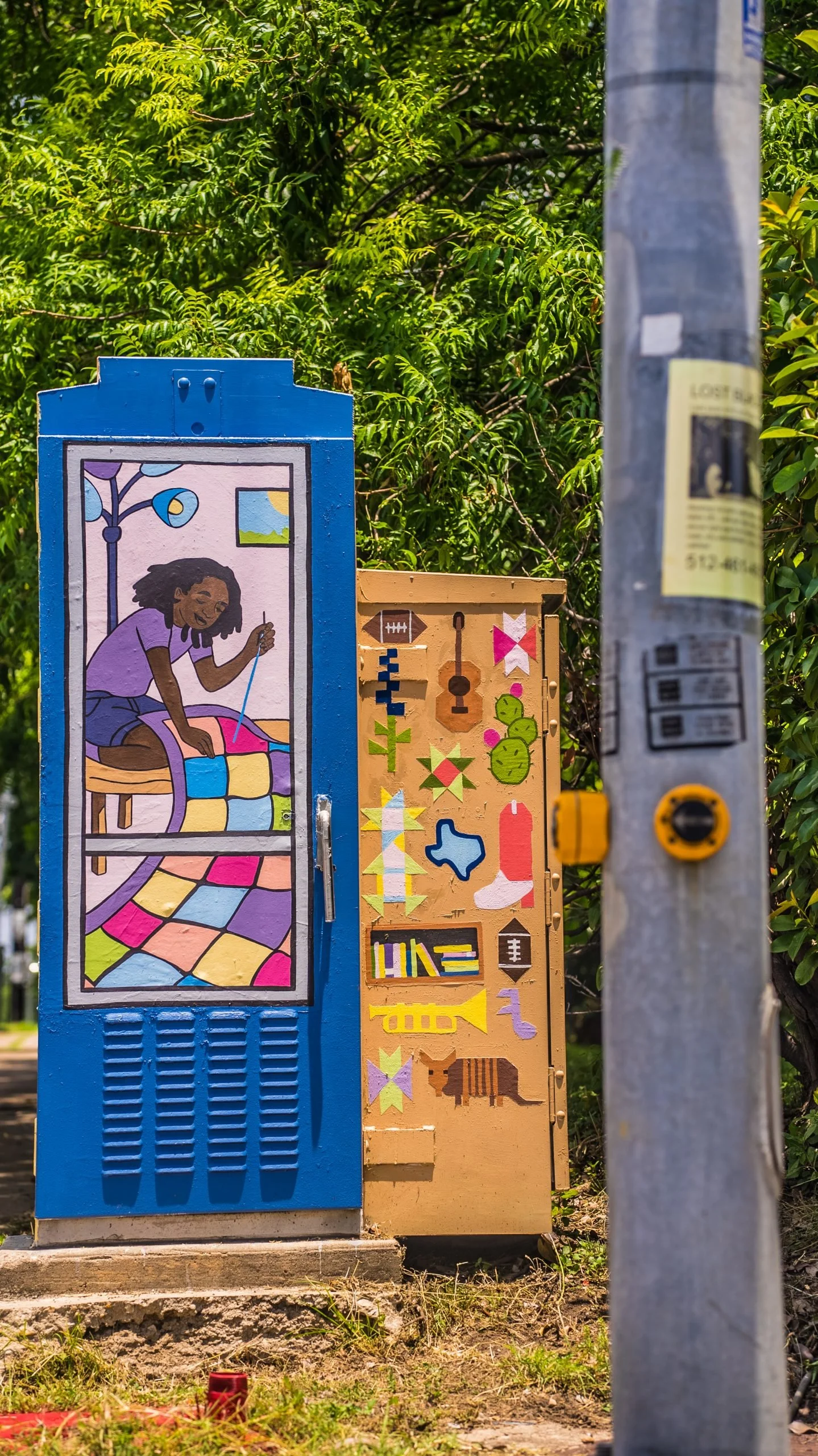 Colorful mural on utility boxes depicting a girl knitting on a pink quilt with a window showing a blue sky and mountains in the background, surrounded by greenery and a gray utility pole.