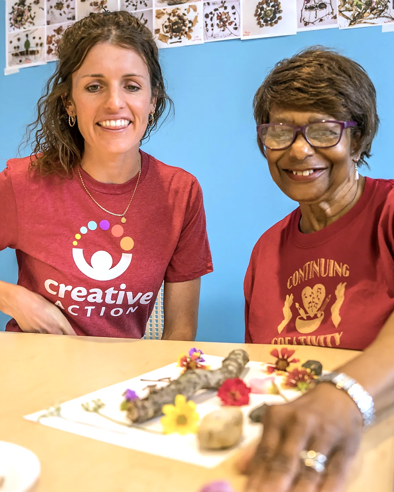 Two women sitting at a table with a display of flowers and sticks, smiling for a photo. They are wearing red shirts with artistic logos, and the background features a blue wall with botanical pictures.