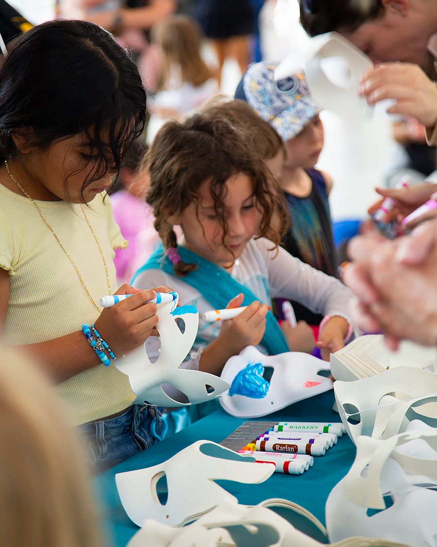 Children decorating paper masks at a craft table with markers and supplies during Community Art Saturday.