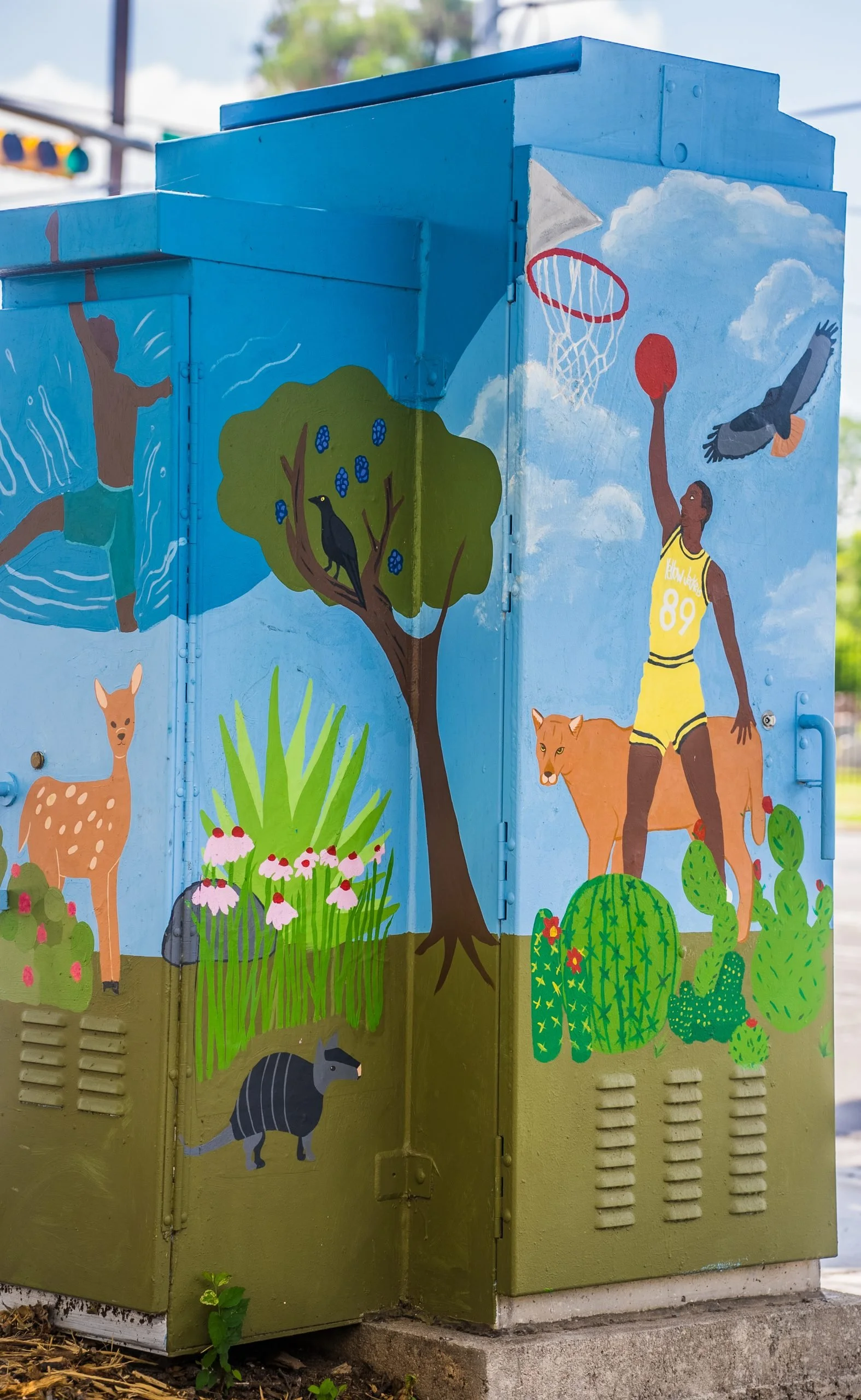 Colorful mural on a utility box depicting various nature and wildlife scenes including a deer, a raccoon, a tree with a bird, a basketball player, cacti, flowers, a bird flying, and areas of sky with clouds.