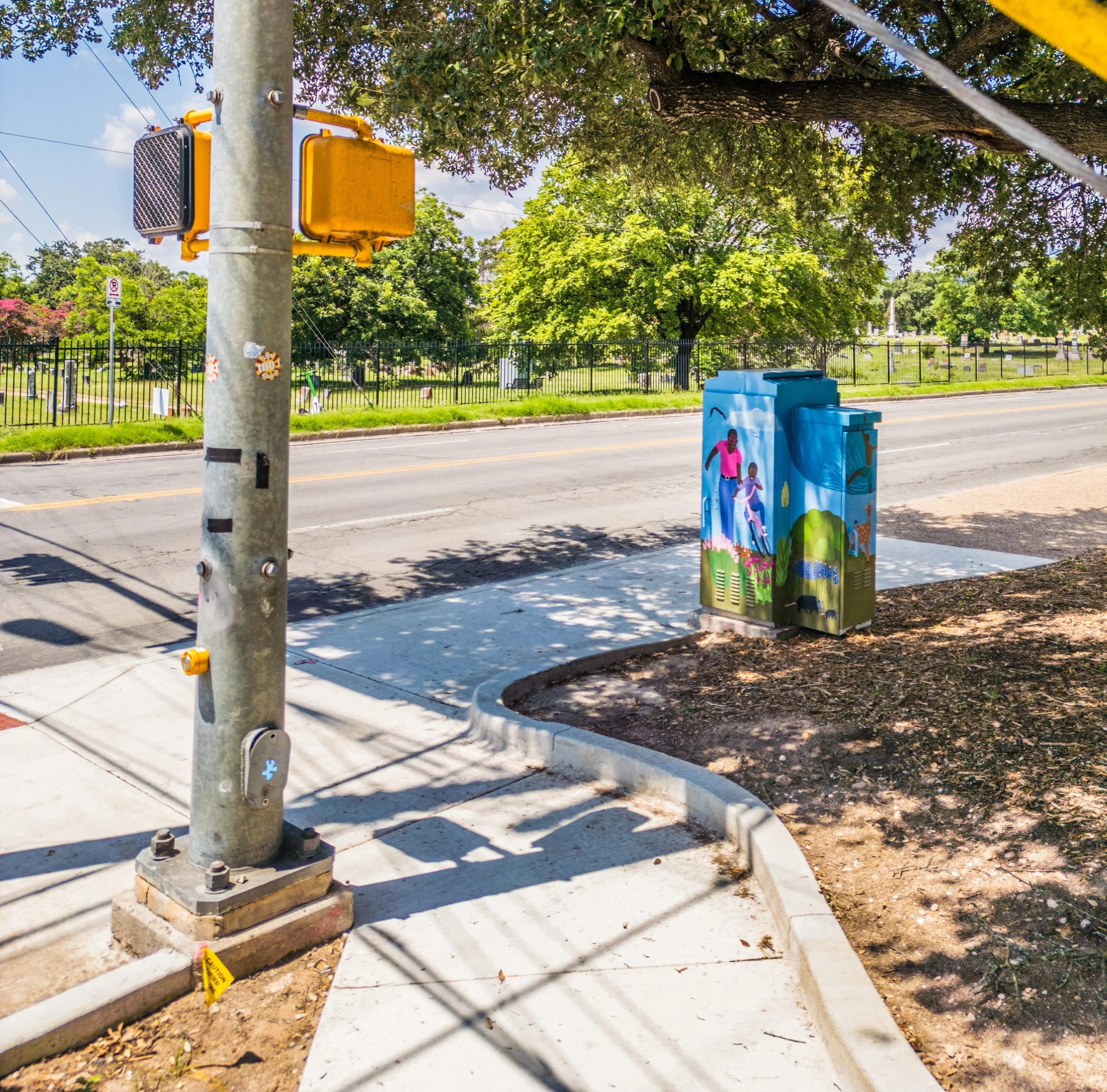 A street scene with a metal utility pole and a yellow electrical box with a large fan near the top. There is a container decorated with children playing and a yellow caution tape on the ground. Trees and a cemetery with headstones are in the background. The sidewalk is shaded by tree branches.