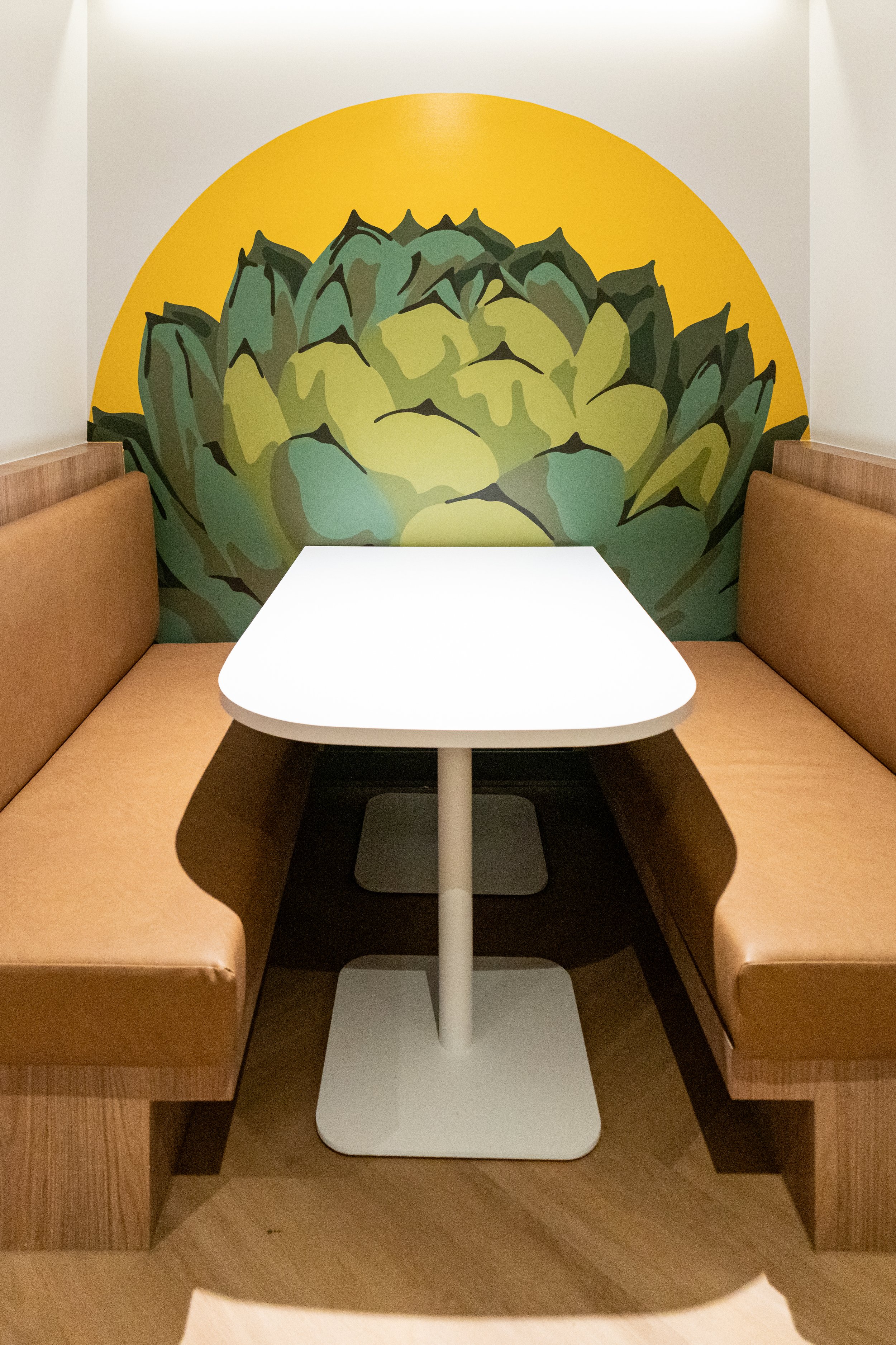 Interior view of a booth with beige cushioned seats on either side of a white table, with a decorative art mural of an artichoke on the yellow semicircular wall.