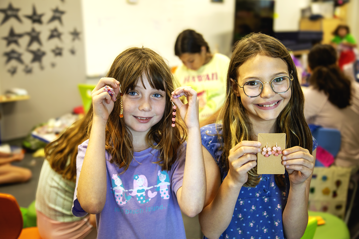 Two young girls smiling and showing jewelry they made, in a classroom with other children and crafting materials in the background.