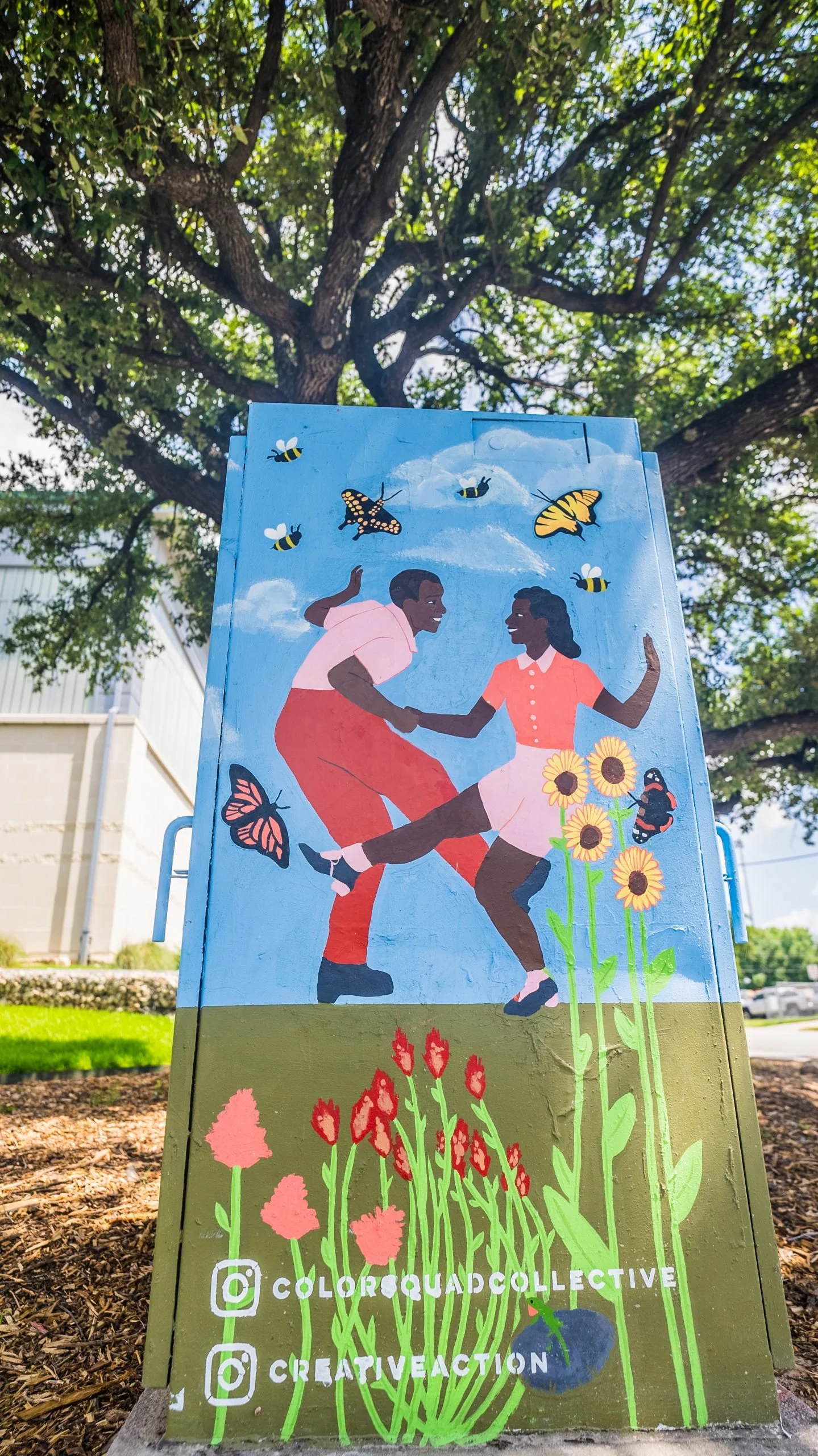 A colorful outdoor mural depicting two women dancing, surrounded by bees, butterflies, sunflowers, and other flowers, with social media handles at the bottom.
