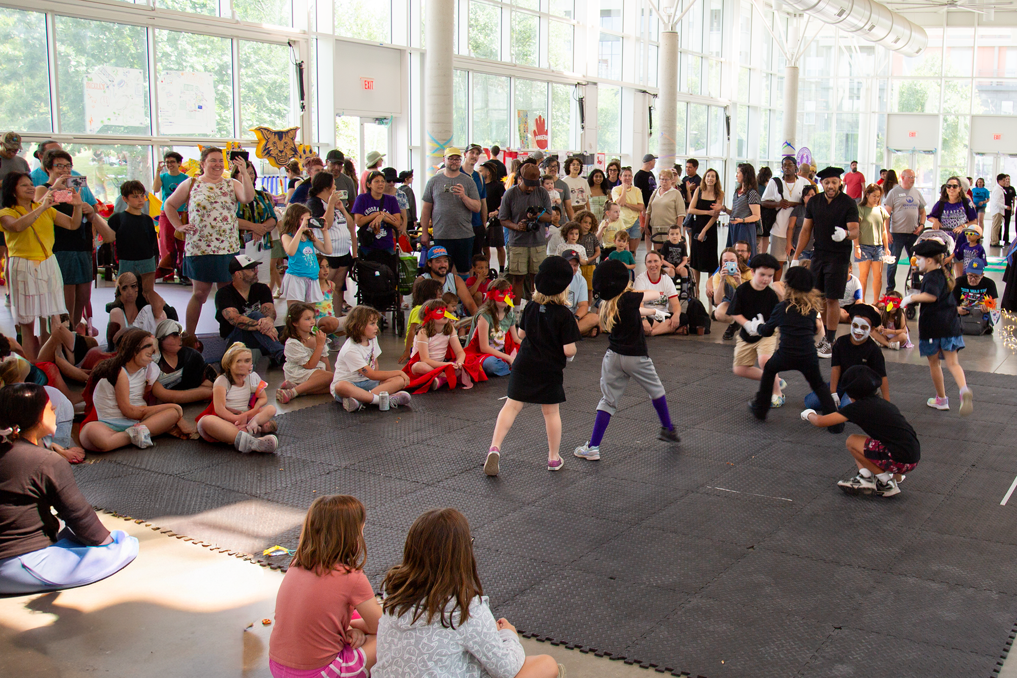 Children and adults watching children perform a dance or skit in an indoor venue with large glass windows and natural daylight.