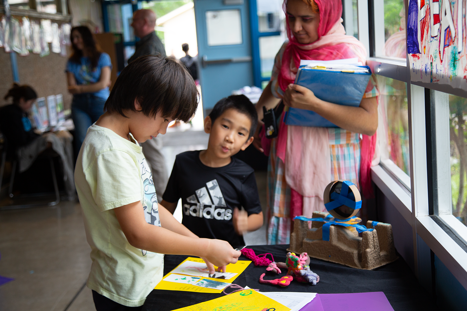 Children and an adult in a classroom looking at projects on a table, with colorful drawings and crafts displayed.