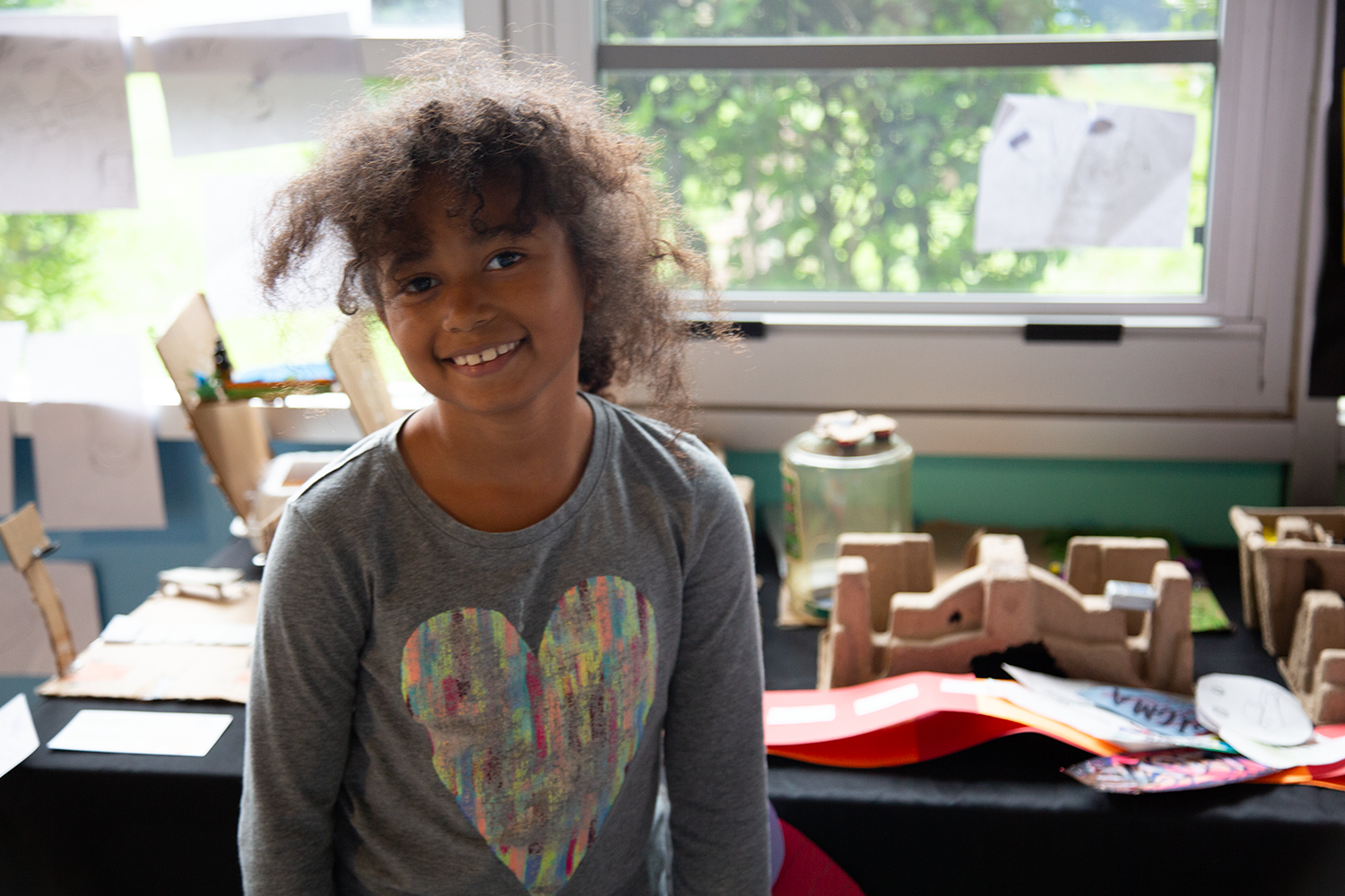 A young girl with curly hair smiling at the camera inside a classroom with art projects on tables in the background during an after school showcase.