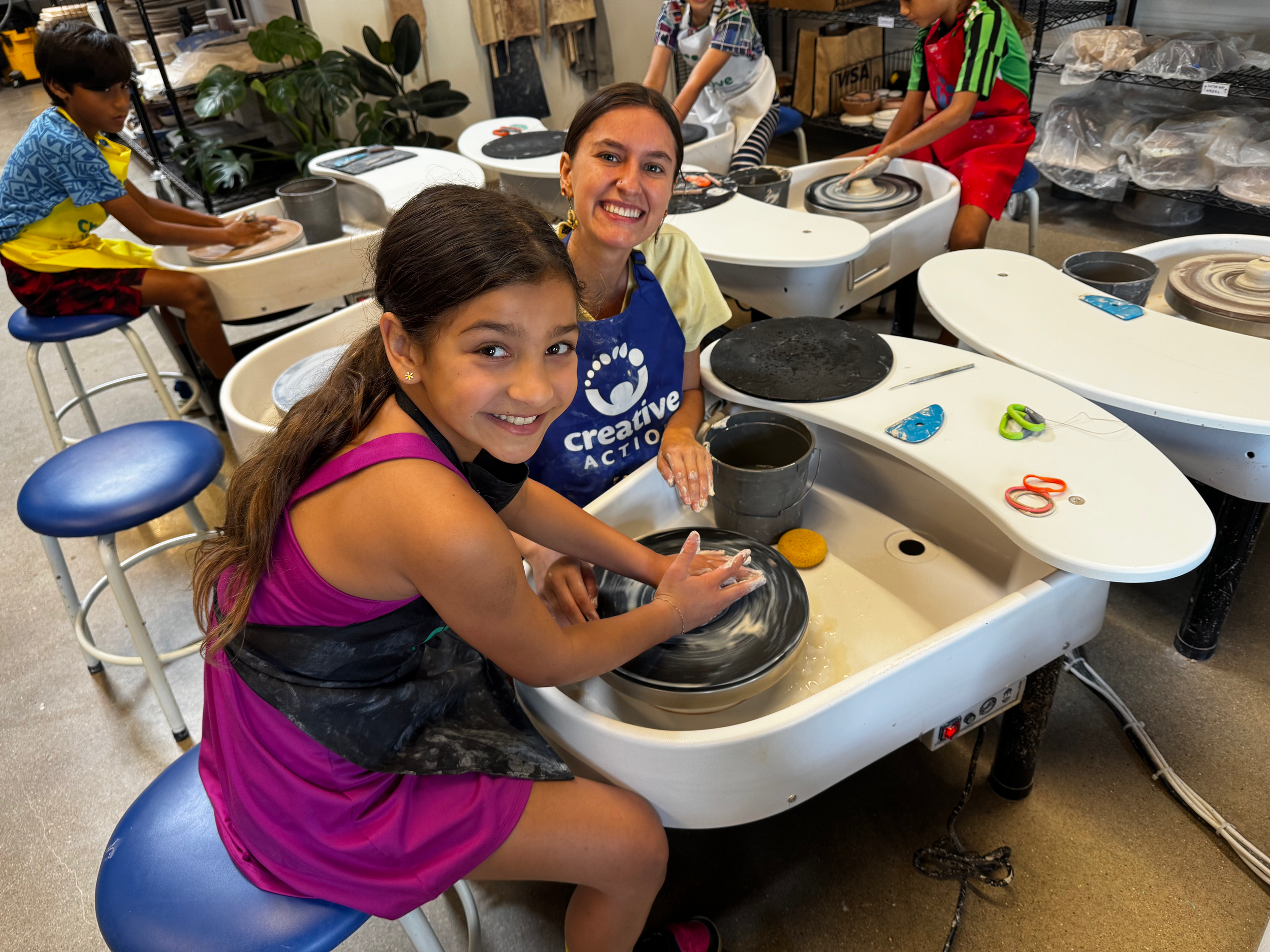 Two girls and two boys making pottery at a pottery wheel workshop. The girls are smiling at the camera, one in a purple tank top and the other in a yellow shirt with a blue apron. The boys are focused on their pottery projects. The workspace includes pottery wheels, tools, chairs, and shelves with supplies.
