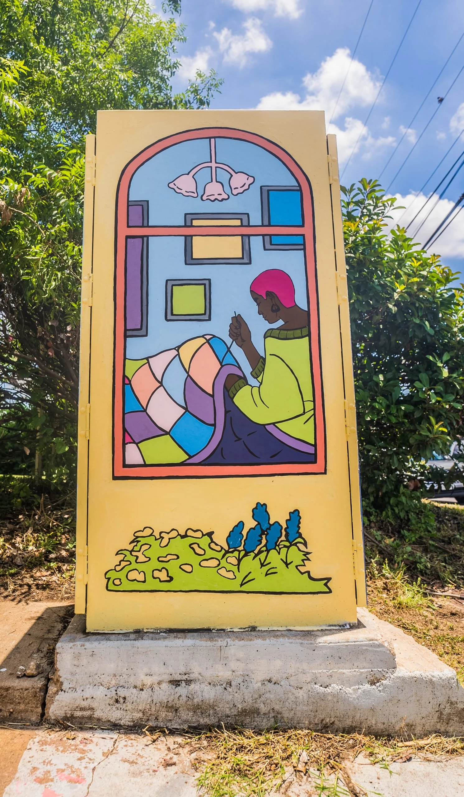 Colorful mural of a woman with pink hair sitting and knitting, inside a window-like frame with a bright blue background, and a plant with yellow flowers at the bottom.
