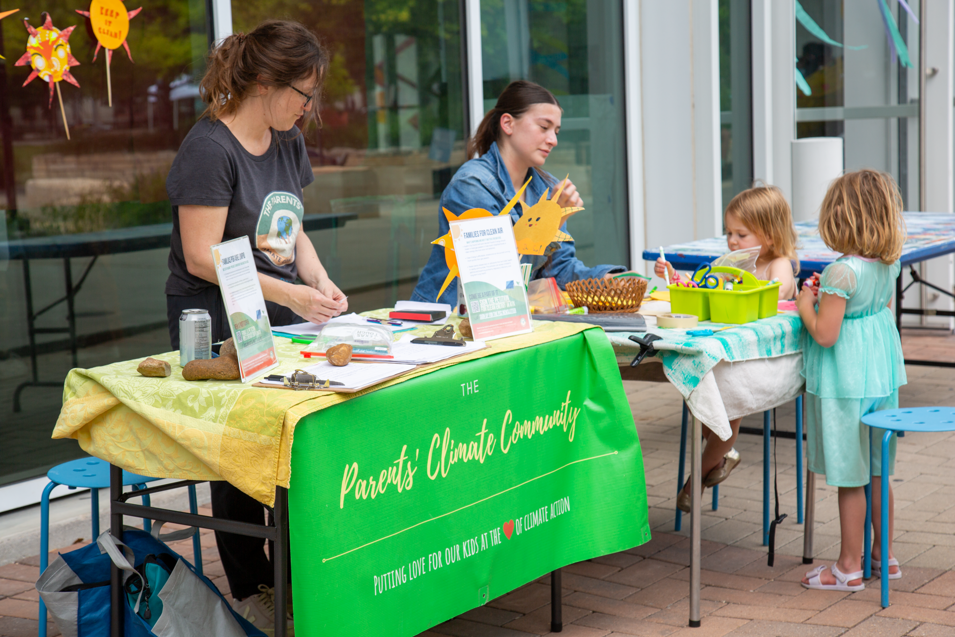 Two women and two young girls at an outdoor event booth promoting climate action. The women are behind a table with informational signs, rocks, and documents, while the girls, one wearing a green dress and the other a light blue dress, stand on the opposite side, engaging with the event materials. The table has a green banner reading "Parents' Climate Community." The background features glass doors and colorful decorations.