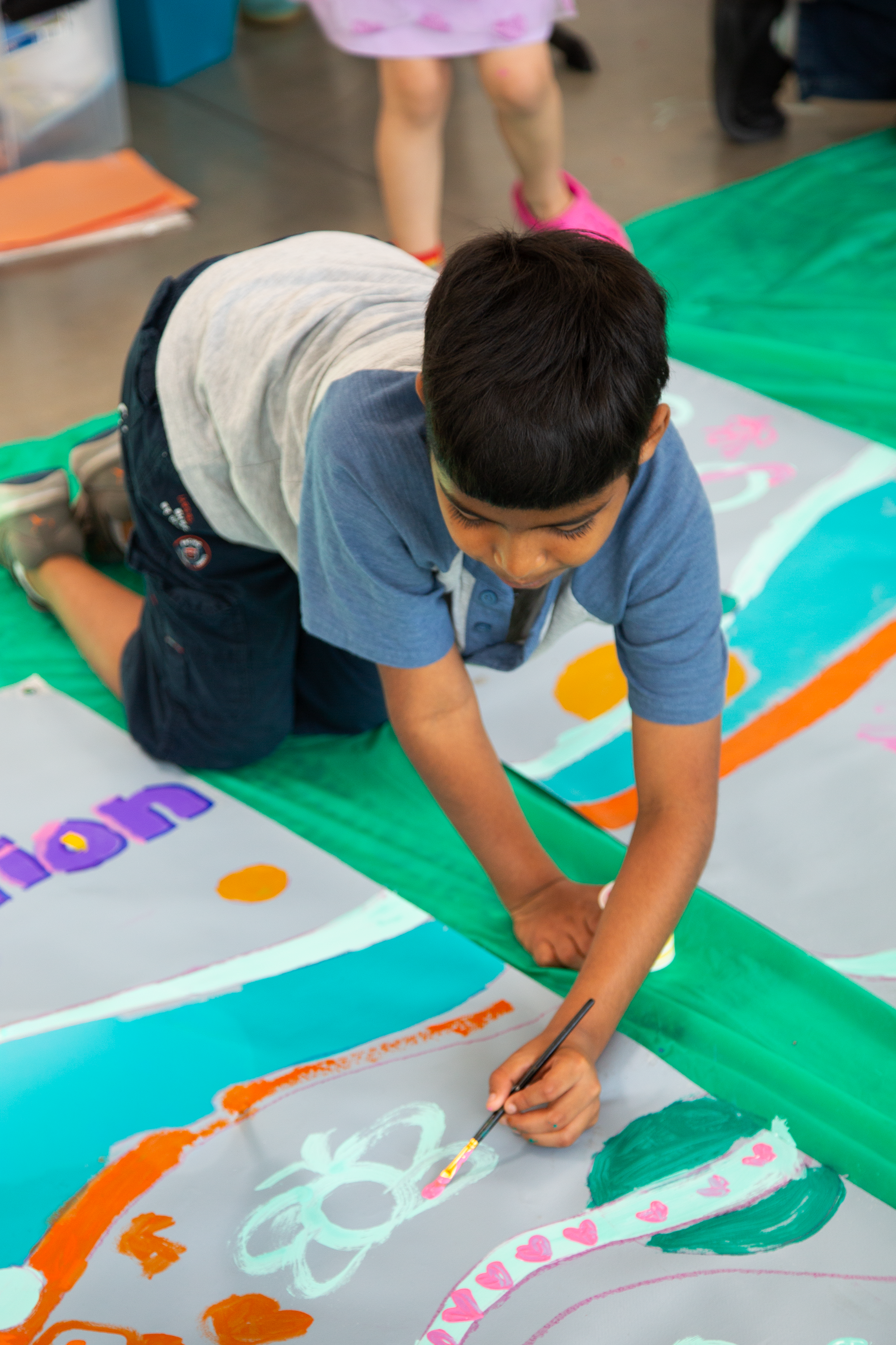 A young boy painting on a large canvas with a paintbrush, colorful rainbow, hearts, and abstract shapes, with other children visible in the background.
