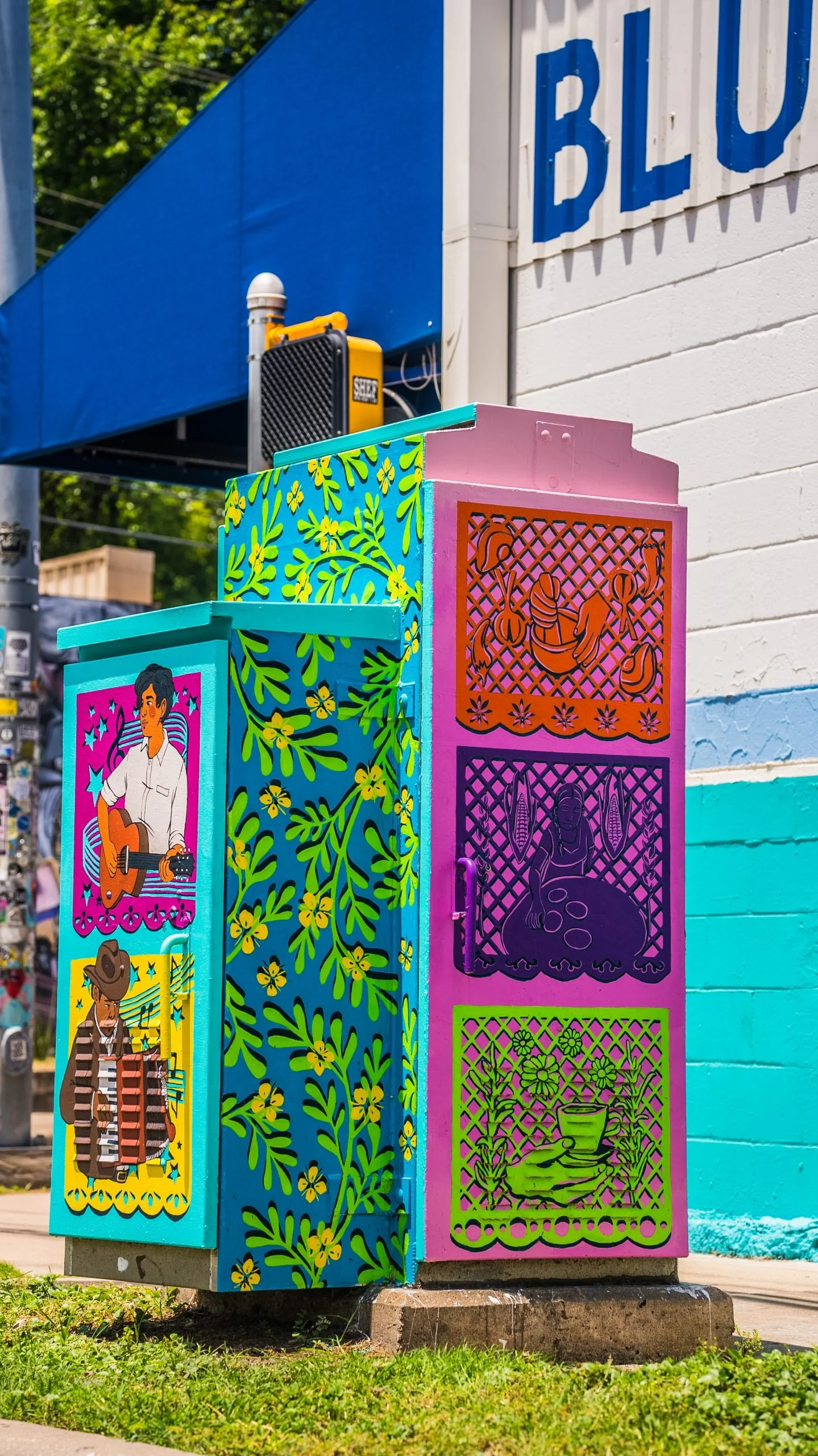 Colorful utility box with vibrant painted panels featuring artistic illustrations of people and nature, situated in front of a white brick building with blue signage and a blue awning.