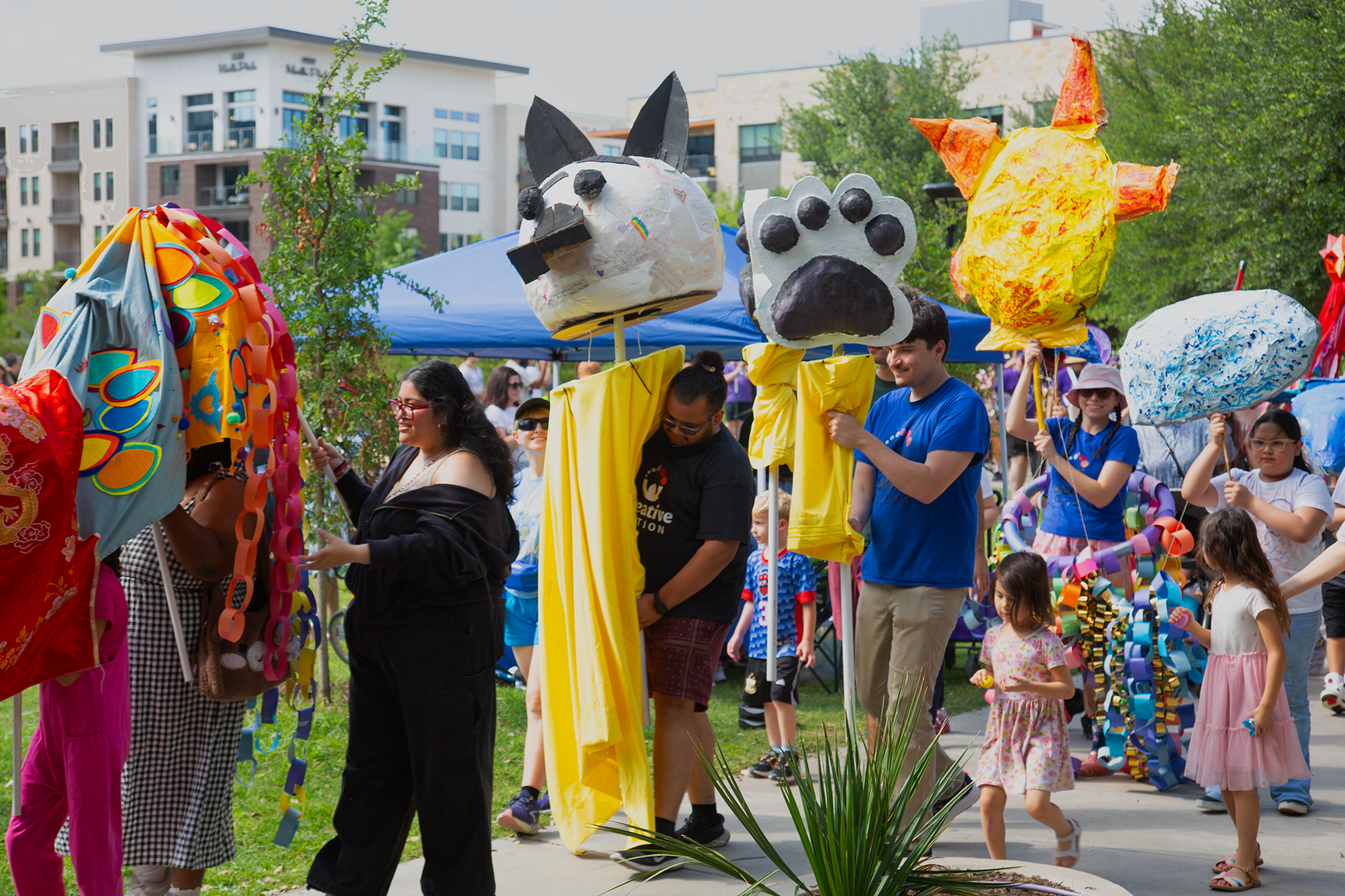 People participating in a parade or festival outdoors, carrying colorful paper-mâché animal and character masks on sticks, with a background of modern residential buildings and trees.