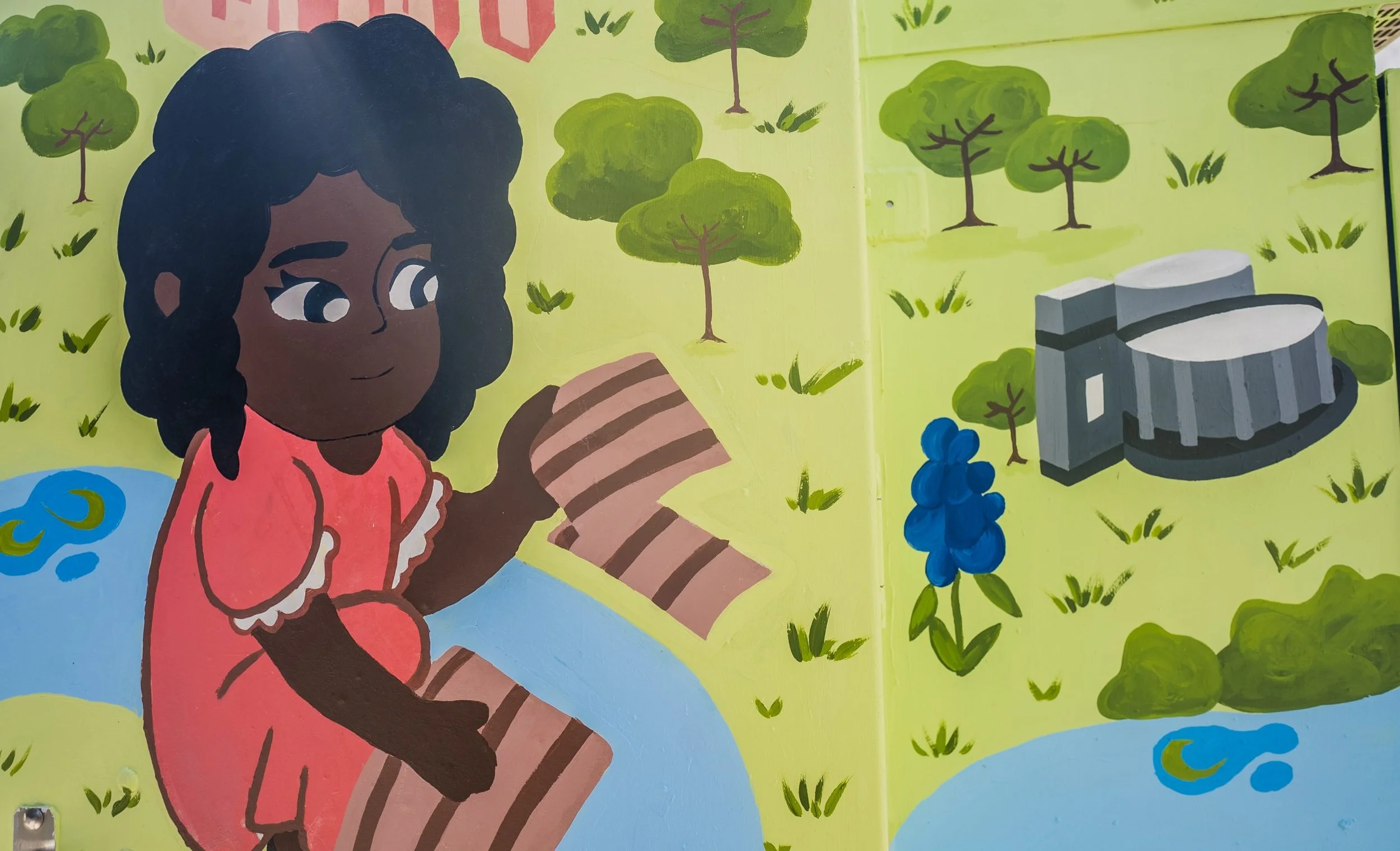 A colorful mural depicting a girl with dark curly hair, wearing a pink dress, sitting on a blue surface and holding a striped brown and beige object. The background shows green trees, grass, and stylized clouds, with a gray stone structure and a bunch of blue grapes.