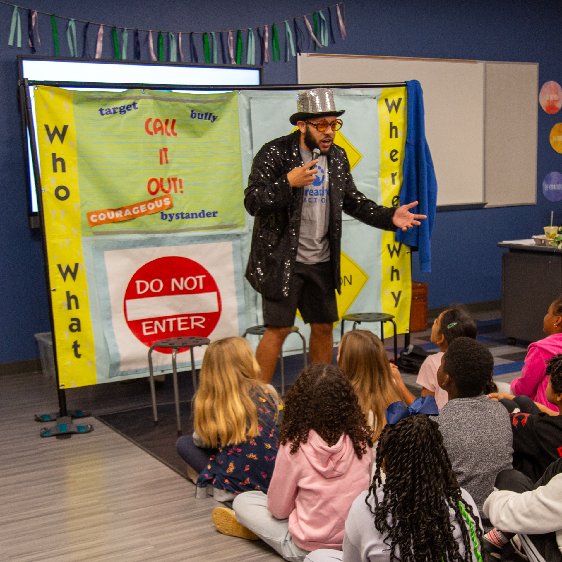 A man dressed as a magician or performer wearing a silver top hat, sunglasses, a black sequined jacket, and shorts, holding a microphone and speaking to a group of children sitting on the floor in a classroom or auditorium. There is a colorful background with banners and signs including a yellow banner with red and blue text, and a white sign with a red circle and