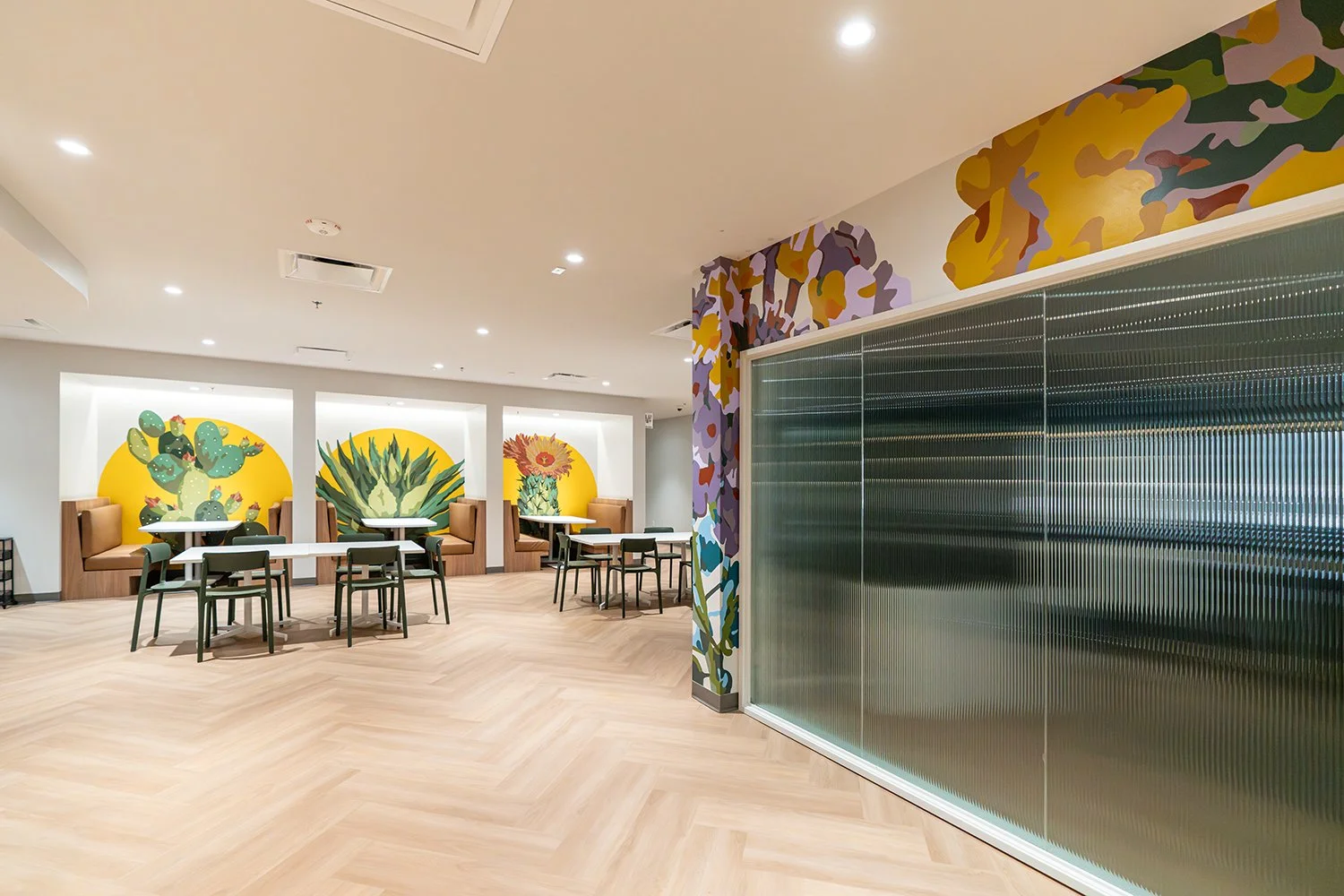 Interior view of a modern dining area with booth seating and tables, colorful cactus and sunflower murals on the wall, a decorative floral wall on the right, and a large frosted glass wall or partition.