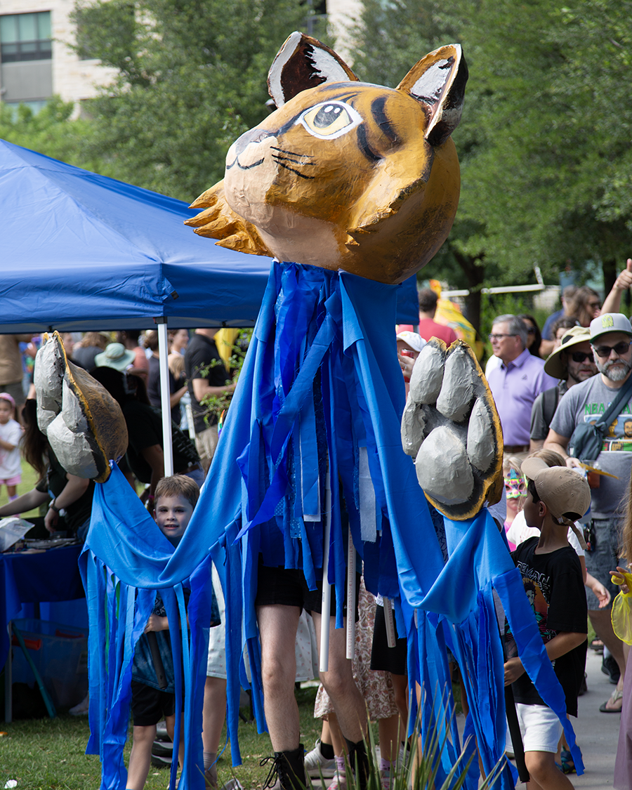 A person wearing a large, colorful cat head costume with blue streamers and paws, standing among a crowd at an outdoor festival or parade.