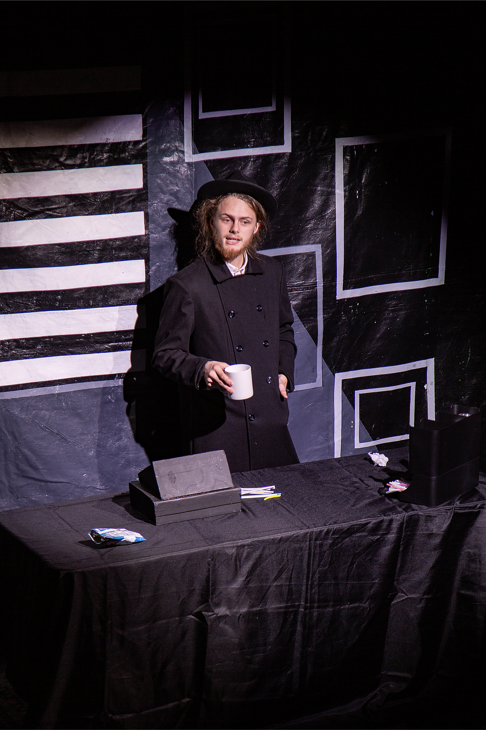A man wearing a black coat and black hat is standing behind a table with various items, holding a white mug. The background is painted with black and white geometric patterns.