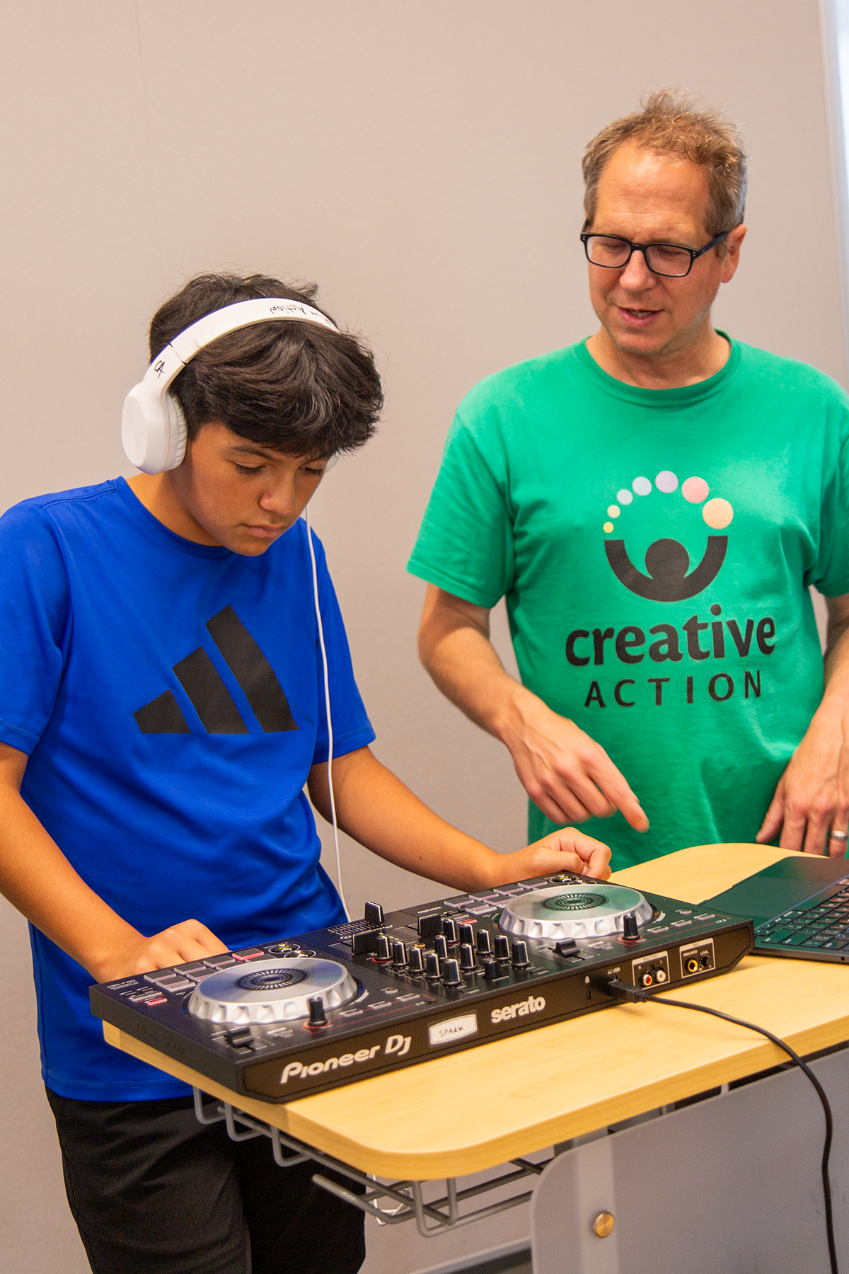 A young person with dark hair wearing a blue Adidas shirt and white headphones DJing on a Pioneer DJ controller, with an adult male in a green shirt standing beside them in a room with a light-colored wall.