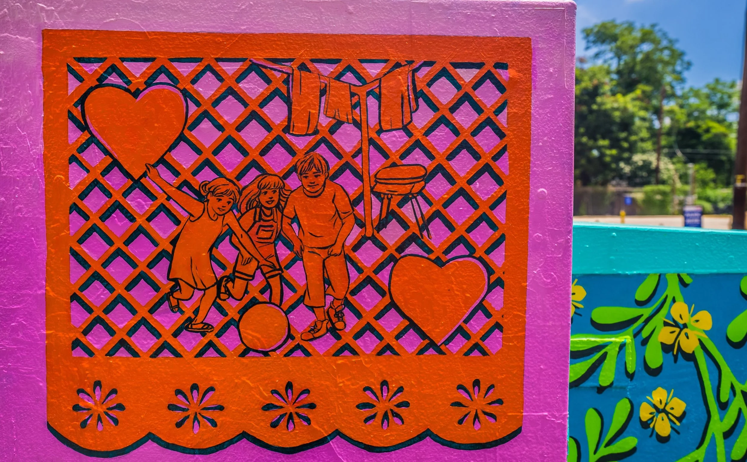 Street art illustration on a purple wall featuring three children playing with a ball, a heart symbol, a flag, and a barbecue grill, framed with floral designs.