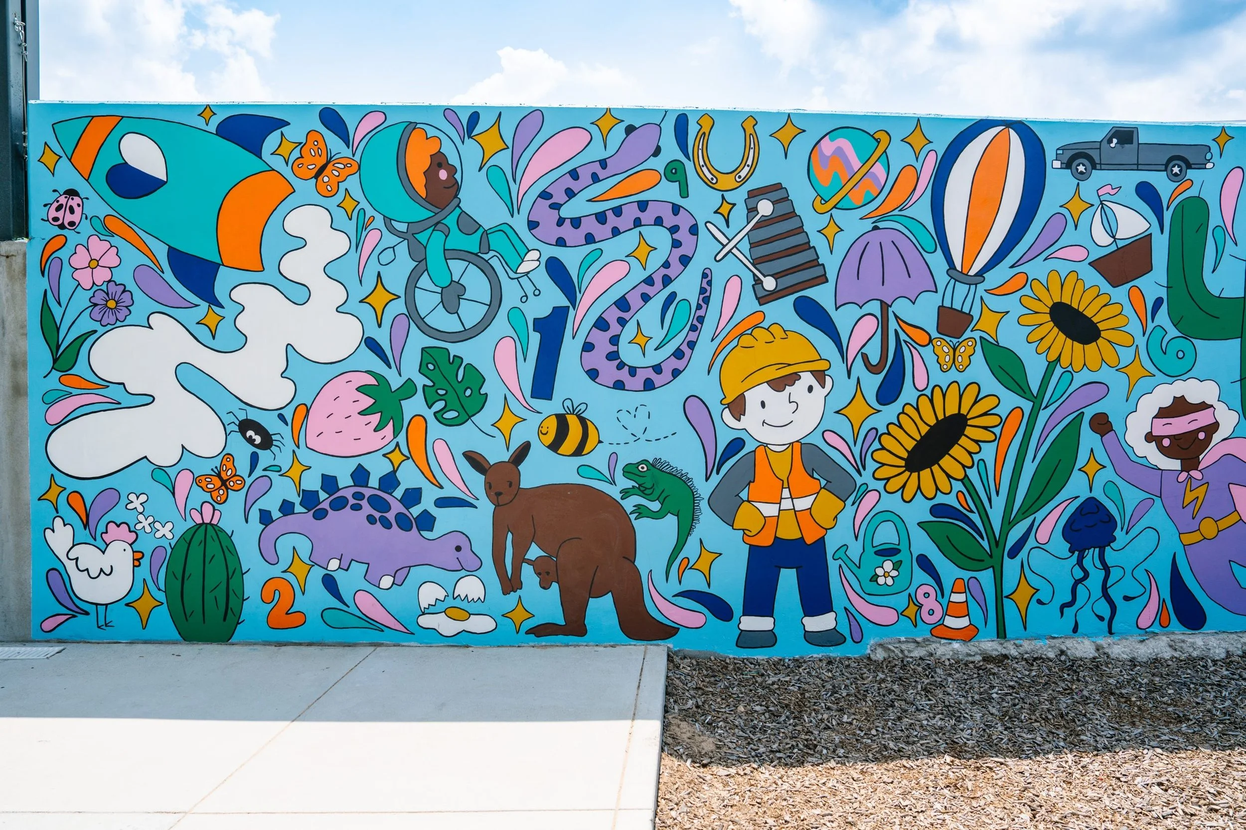 Colorful mural featuring children, animals, and whimsical elements like butterflies, flowers, clouds, a spaceship, a UFO, and a truck on a bright blue background.