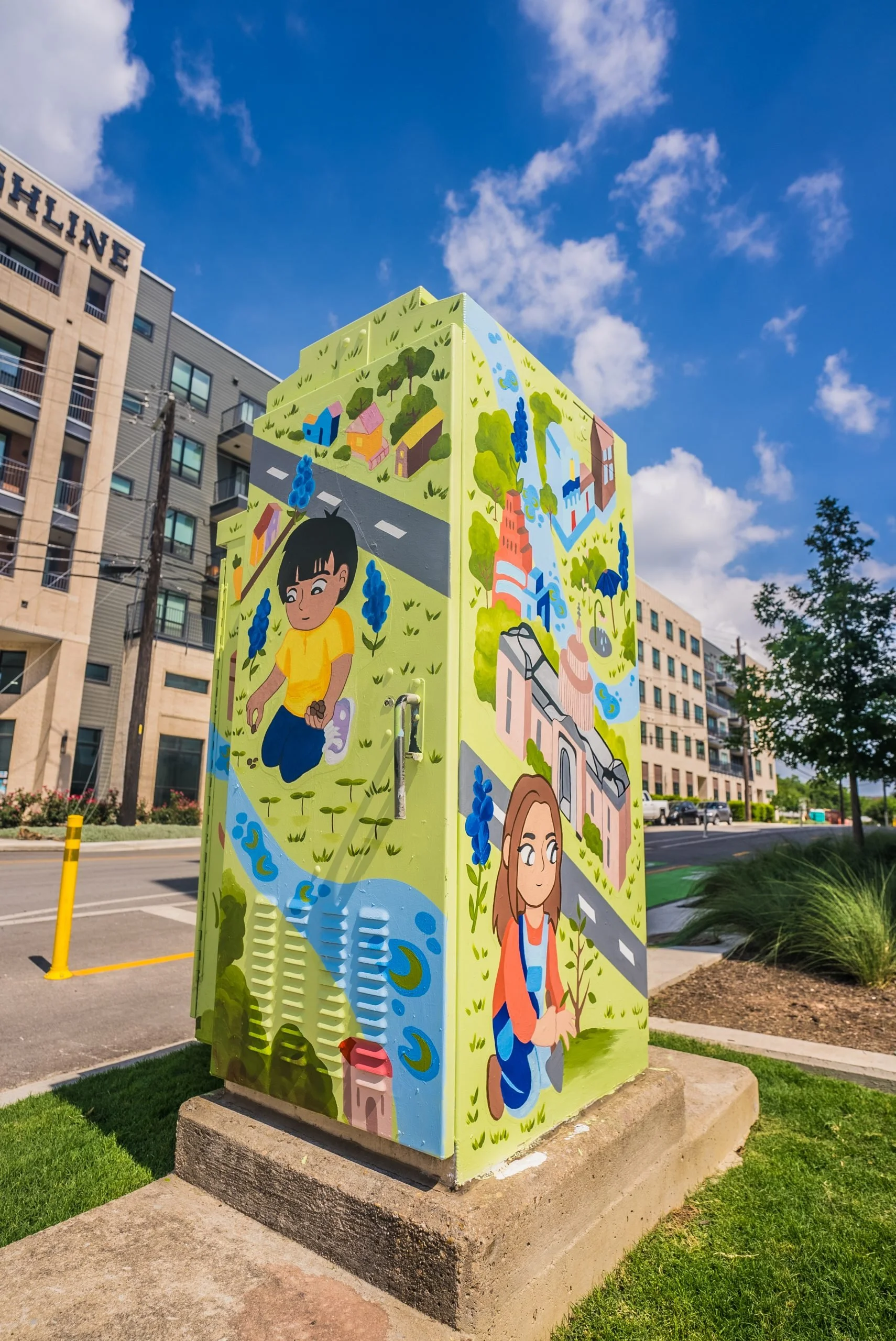 Colorful mural on a utility box depicting children playing outdoors, with trees, a river, and a blue sky with clouds, set in an urban environment with buildings and a street in the background.