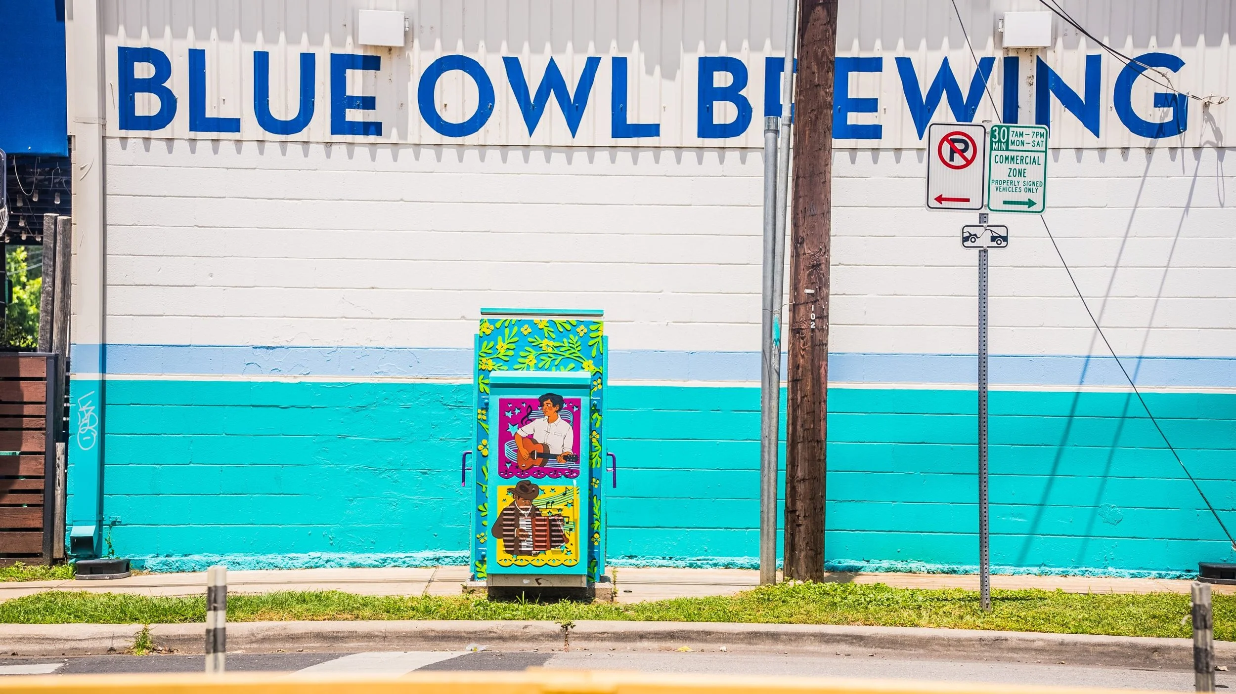 A wall painted with light and dark blue horizontal stripes with the word 'BLUEOWL' in large blue letters. In front, there is a colorful mural of a person playing a guitar and another person wearing a hat. There are street signs indicating parking regulations and a utility pole with wires.